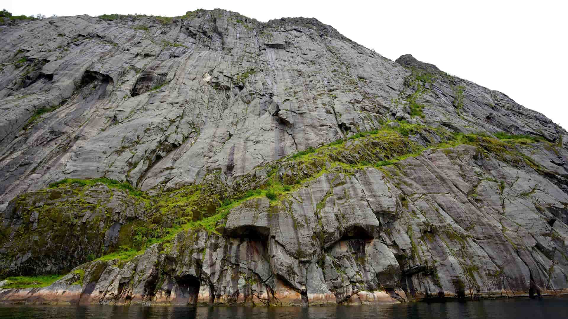 A low-angle shot of a massive, imposing cliff face rising dramatically from the dark water, with patches of moss and greenery clinging to its textured, gray rock surface.