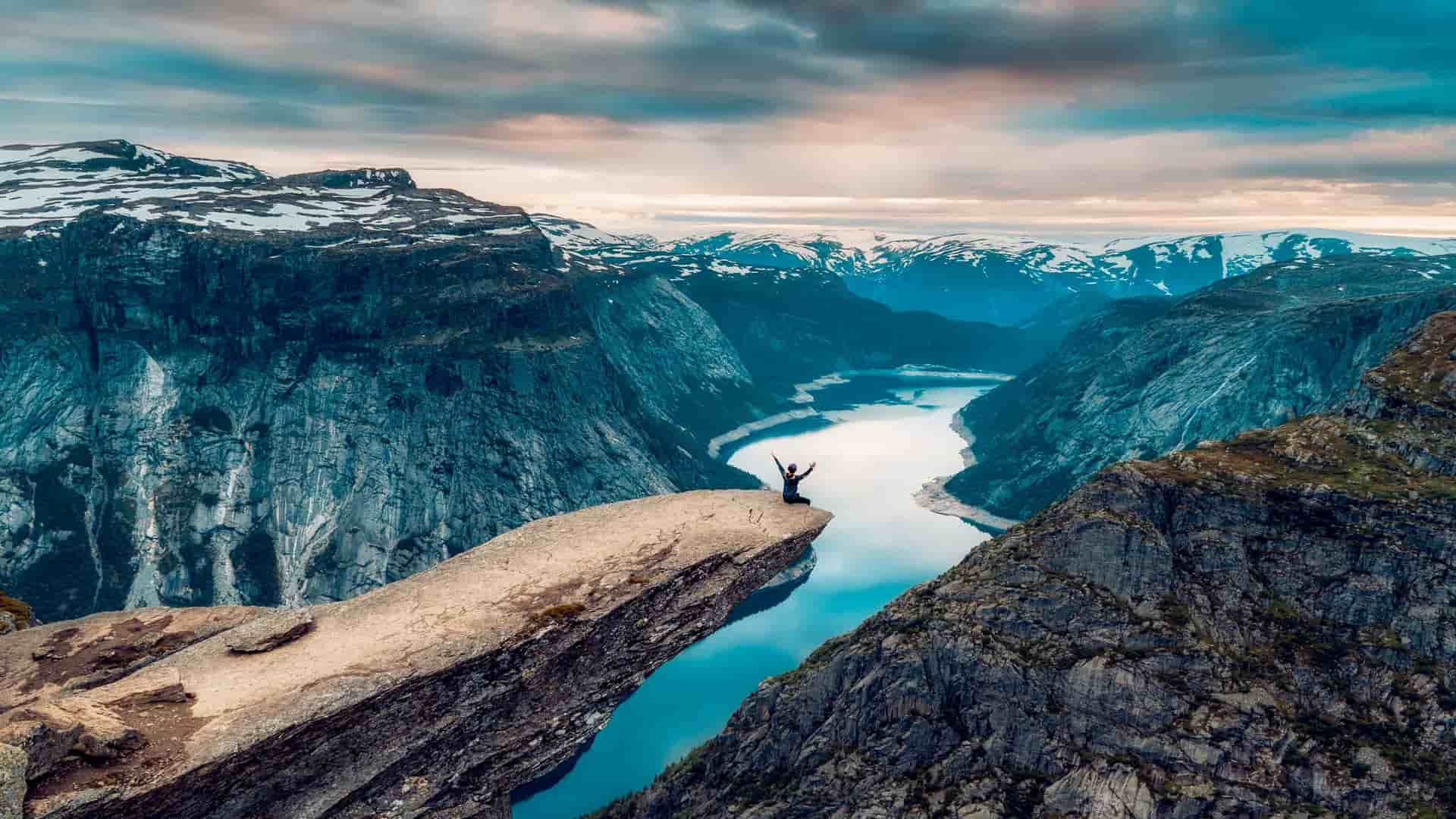 A wide-angle landscape of a person with their arms raised in triumph on the dramatic rock formation of Trolltunga, a major Norwegian landmark, overlooking a glacial lake.
