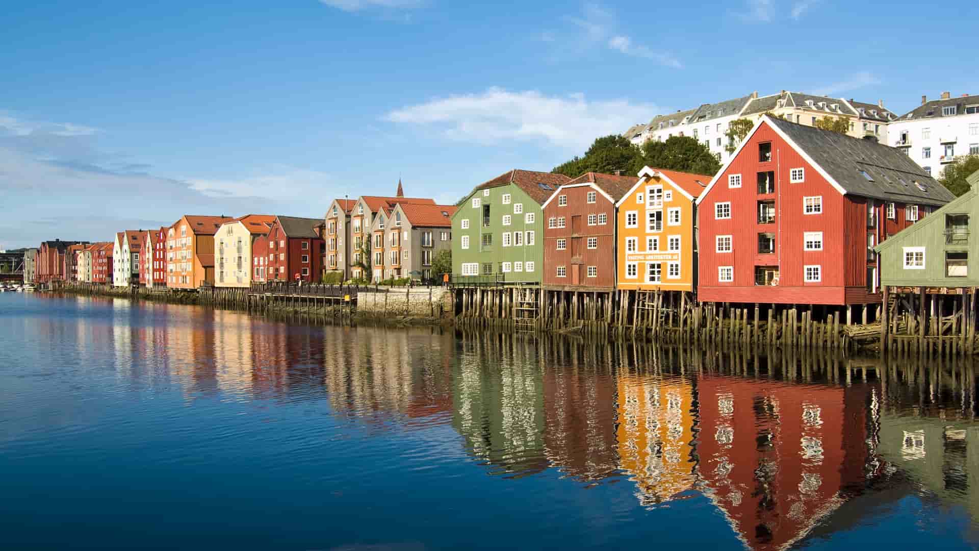 A scenic view of historic, colorful wooden warehouses built on stilts along the Nidelva River in Trondheim, Norway, with their reflections visible in the calm water.