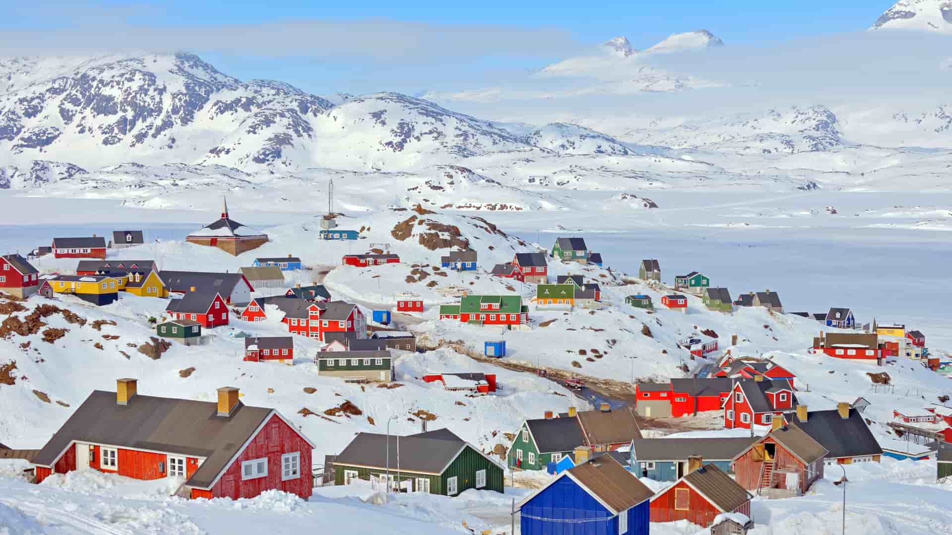 A panoramic view of the colorful houses of Uummannaq, Greenland, nestled on a snowy hillside overlooking a frozen fjord with majestic snow-covered mountains in the background.