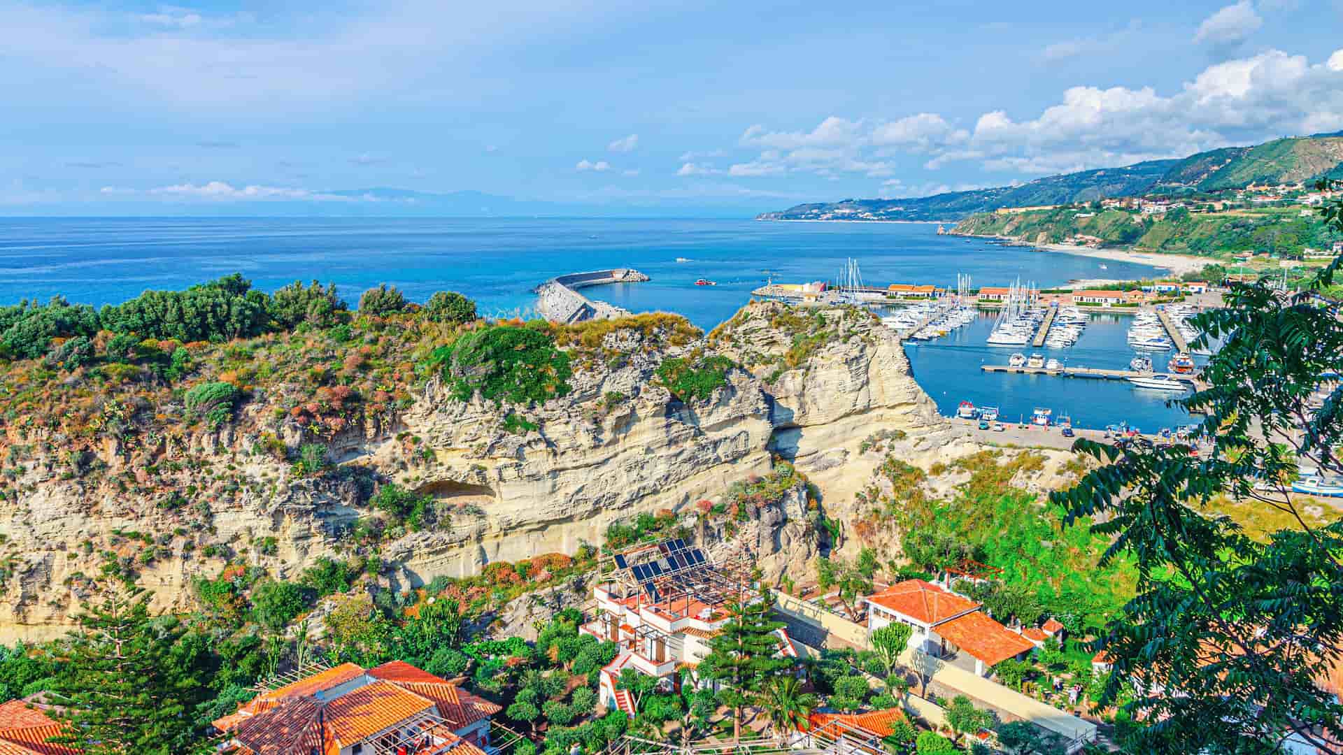 A high-angle view of the scenic coastline of Vibo Valentia Marina in Calabria, Italy, with cliffs, a marina filled with boats, and a beautiful beach, all against the backdrop of the blue Tyrrhenian Sea.