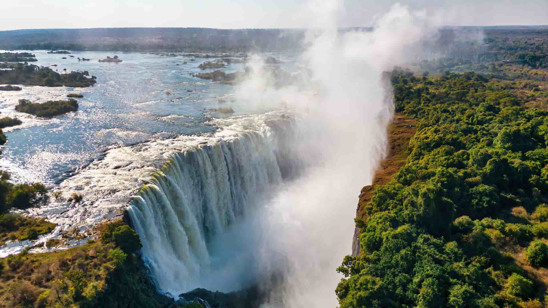 An aerial view of the majestic Victoria Falls, with a massive curtain of water plunging into a gorge, creating a cloud of mist over the lush green forest in Zimbabwe.