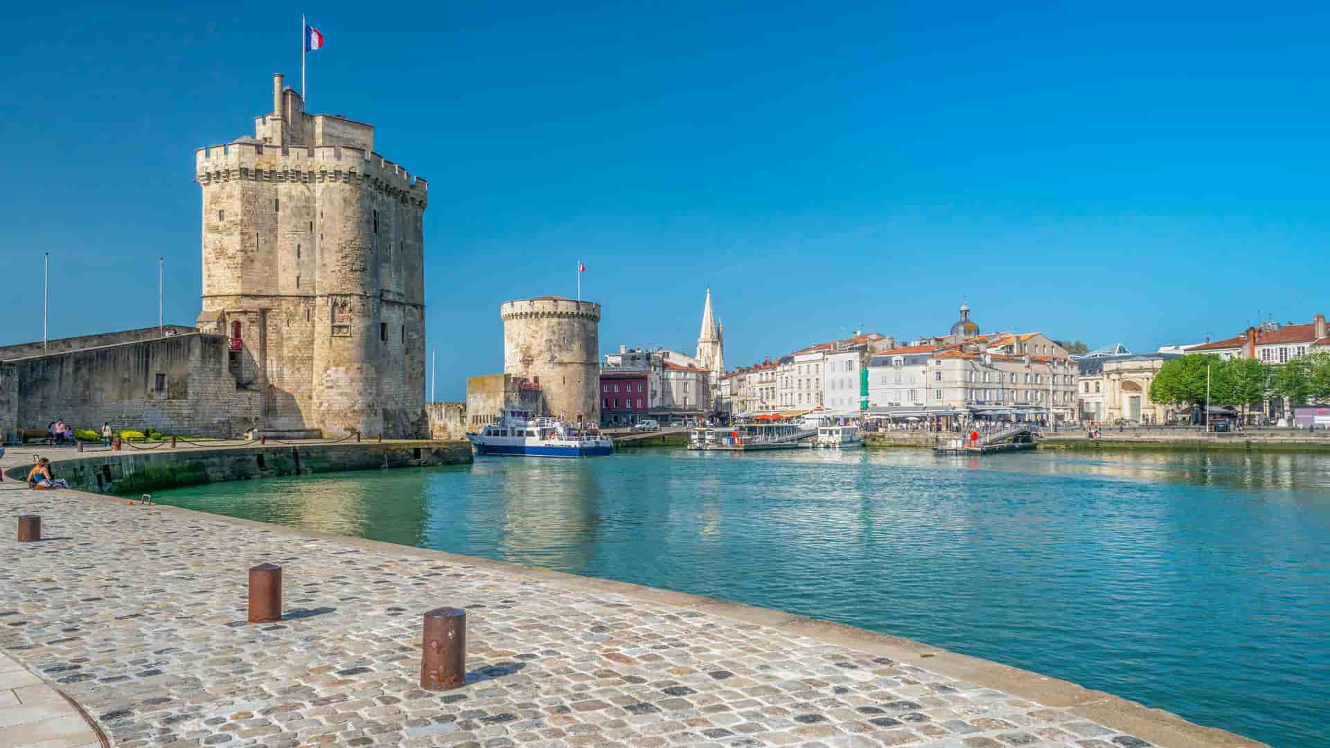 A vibrant harborside view of the historic medieval Towers of La Rochelle, a major landmark, with a cobblestone quay and boats floating in the calm water on a sunny day.