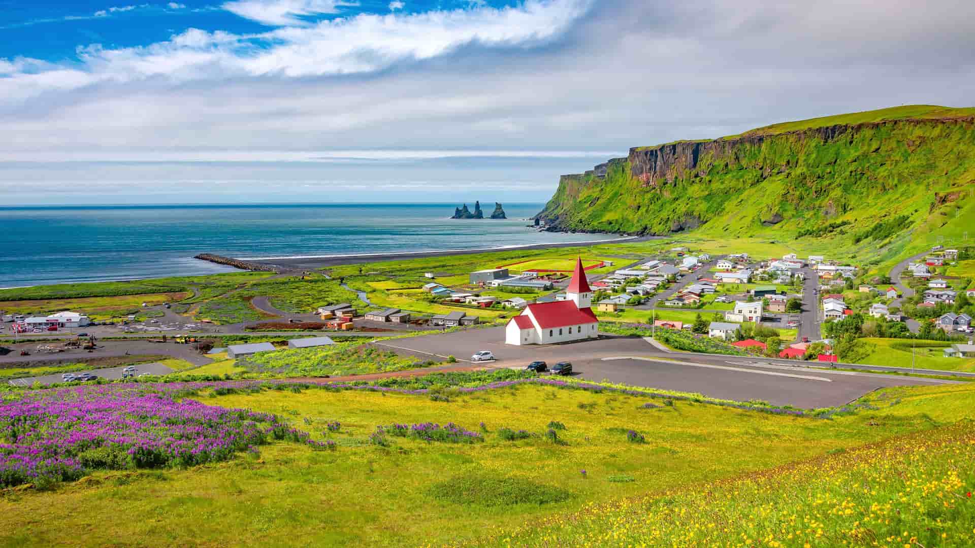 A beautiful summer landscape of the coastal town of Vik, Iceland, with its iconic red-roofed church and a black sand beach against a backdrop of green cliffs.