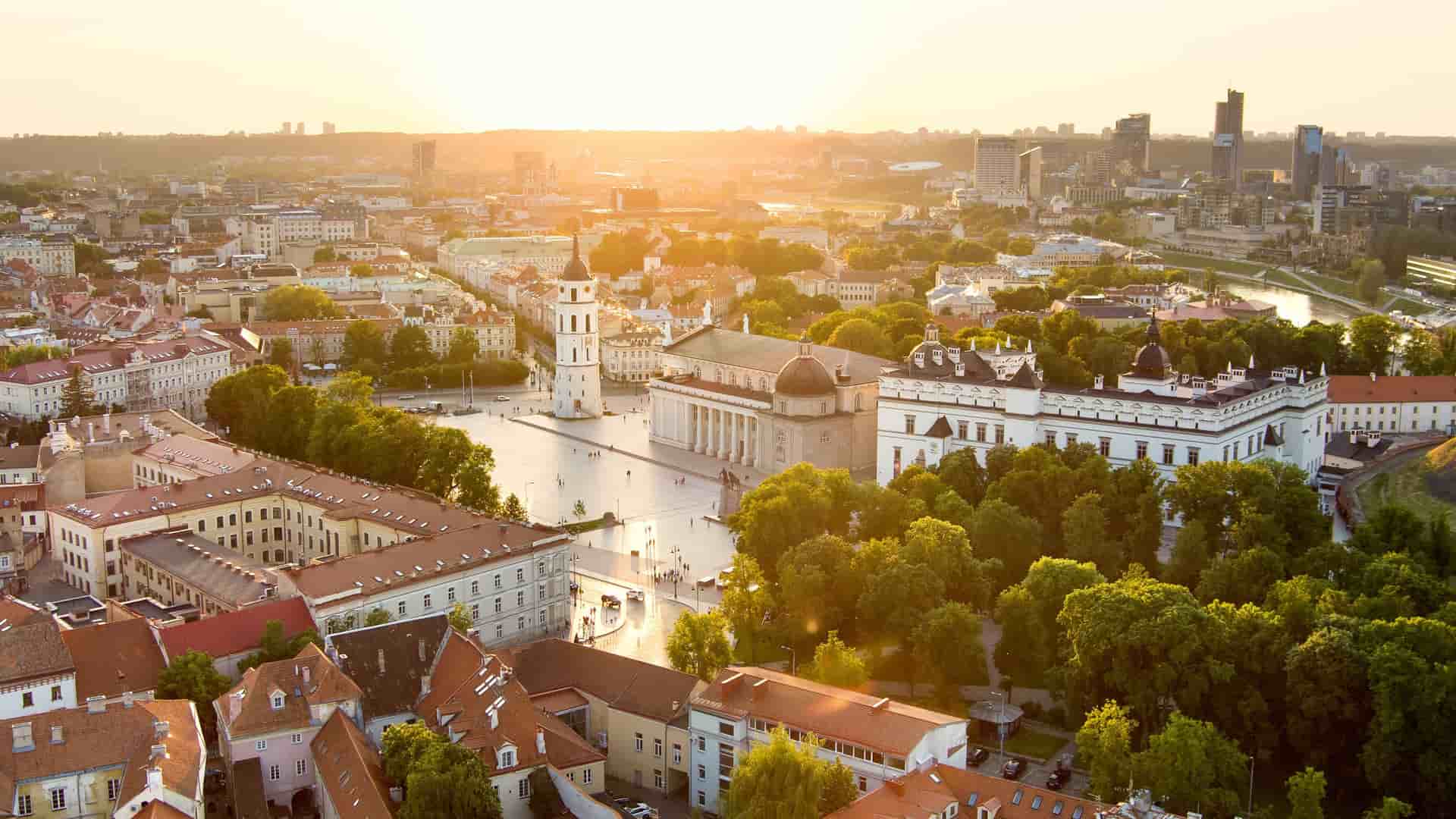 "An aerial view of Vilnius, Lithuania, at sunset, showcasing the historic Vilnius Cathedral and its prominent bell tower in Cathedral Square, with the modern cityscape and the Neris River in the background.  "