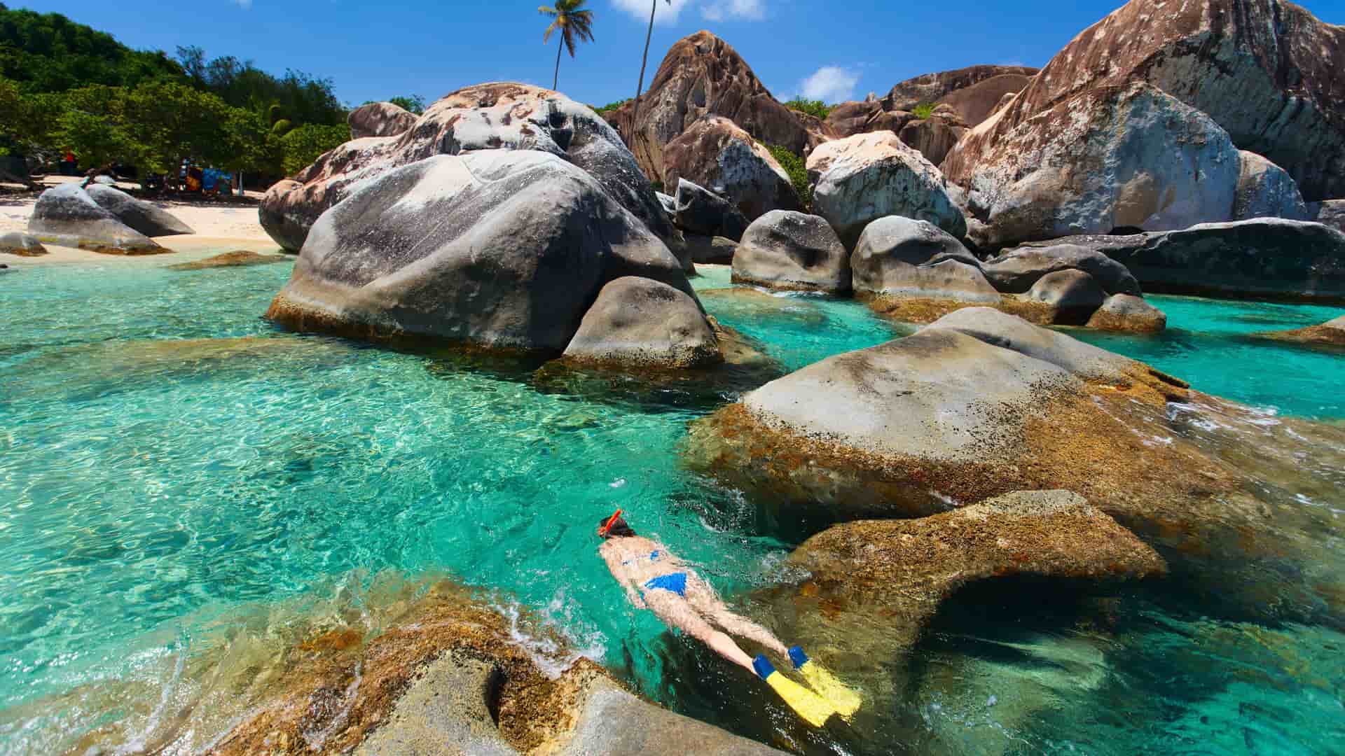 A snorkeler swims in the turquoise waters of the Baths National Park on Virgin Gorda, surrounded by massive granite boulders and palm trees.