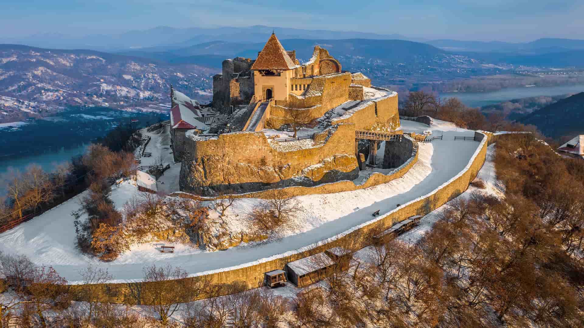An aerial view of the snow-covered ruins of Visegrád Castle in Hungary, with the Danube River visible below and the surrounding mountains in the background.