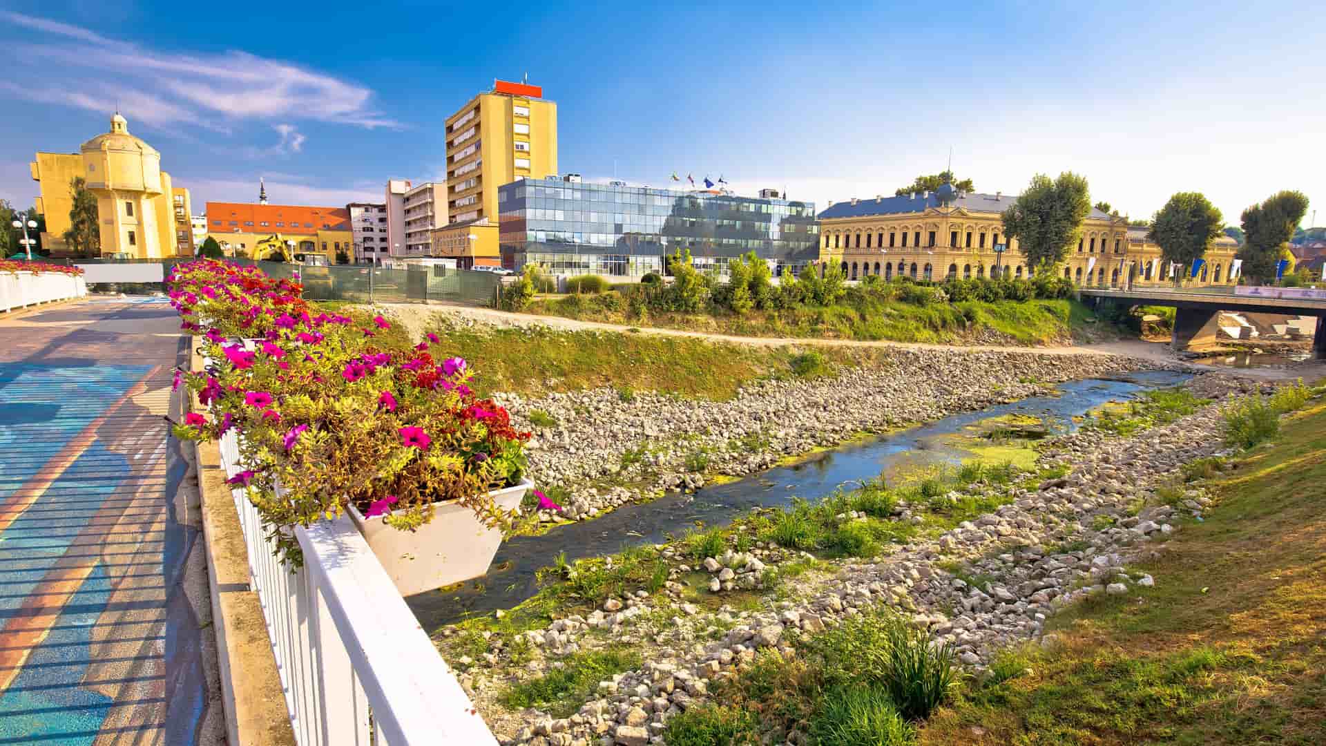 A vibrant riverfront view in Vukovar, Croatia, showcasing the old and modern cityscape with the Duna River, colorful flowers on a bridge, and a mix of historic and new buildings in the background.
