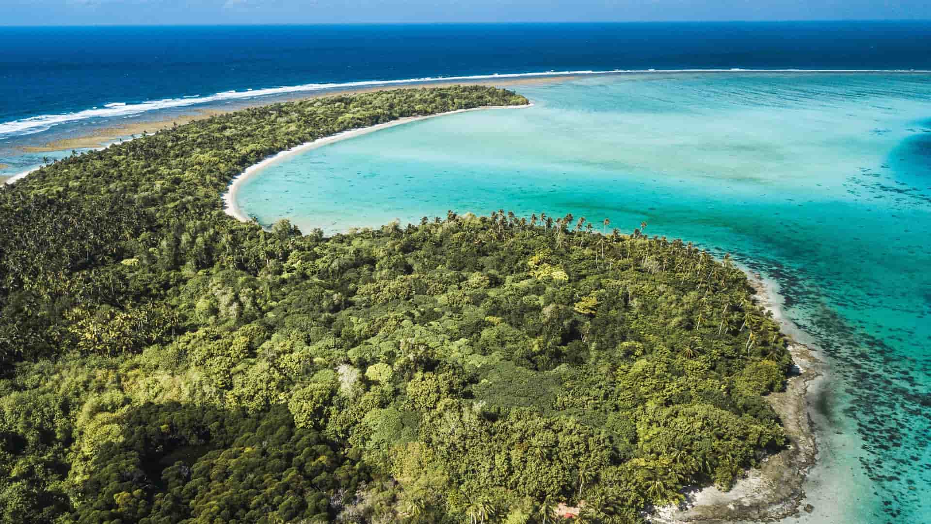 An aerial view of a crescent-shaped, heavily forested tropical island and its surrounding turquoise lagoon on Wallis Island.