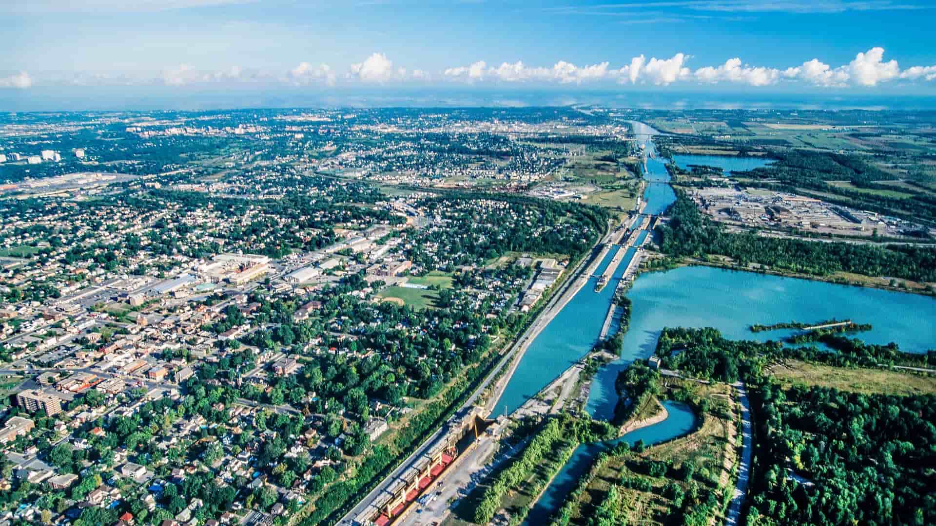 An aerial view of a cargo ship navigating the Welland Canal, with the locks and surrounding parkland, separating the city and Lake Ontario from the sprawling suburbs and industrial areas.