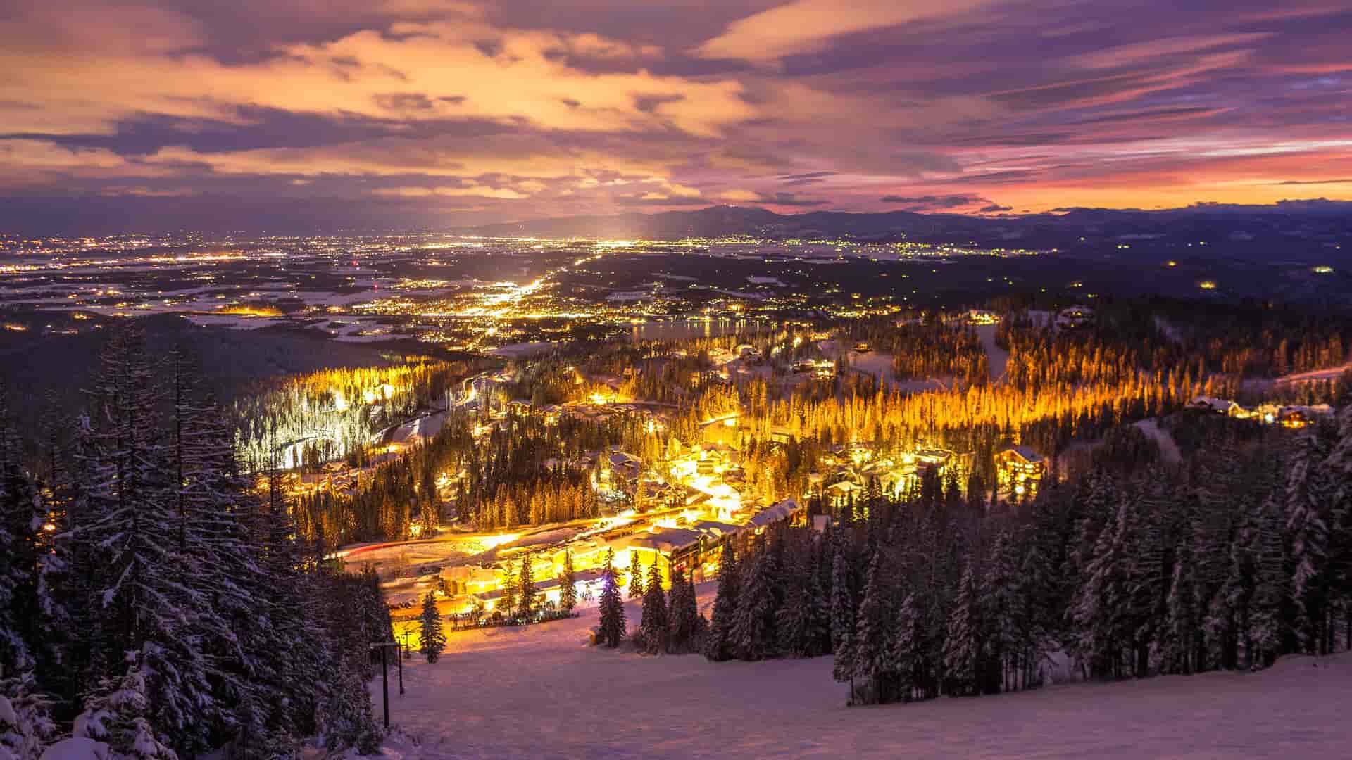 "A panoramic nighttime view of the Whitefish Mountain Ski Resort in Montana, with bright lights illuminating the slopes and surrounding town, under a vibrant sunset sky.  "