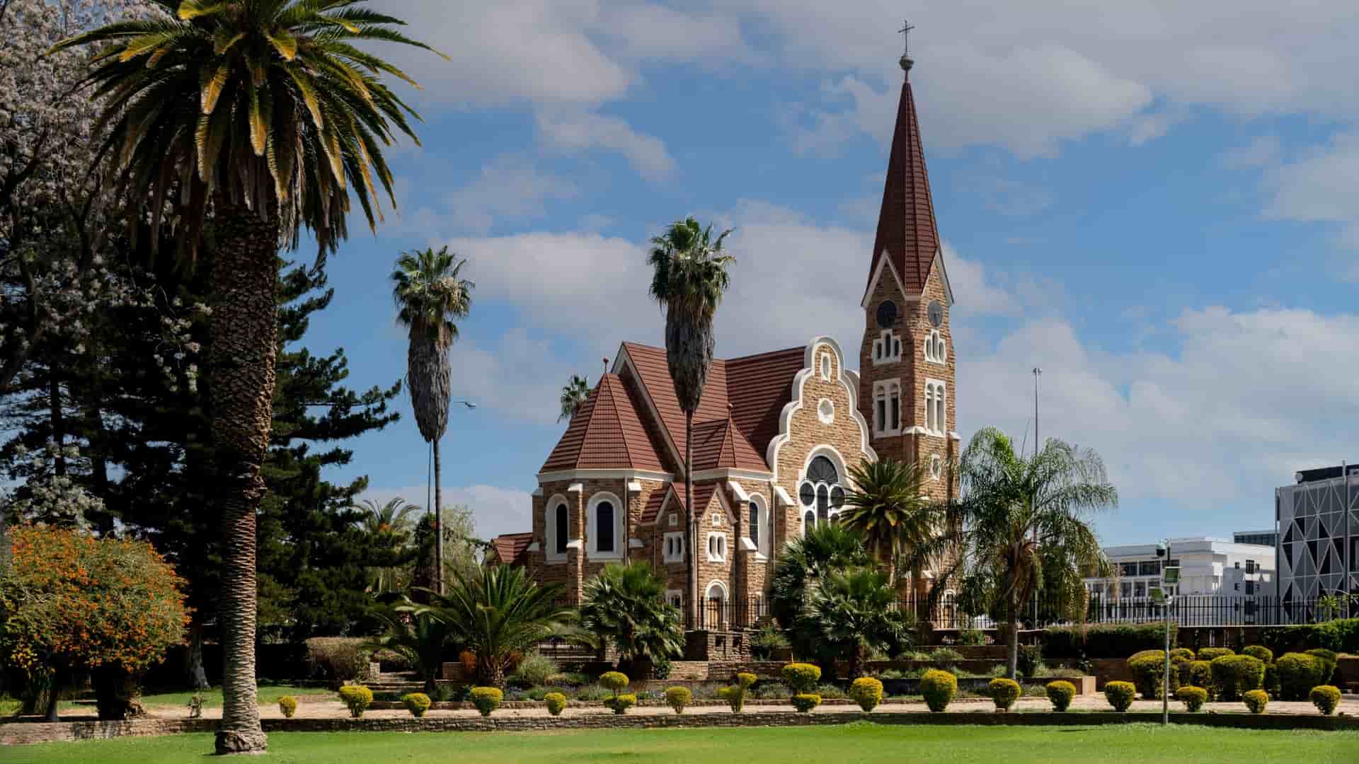 The Christ Church is a neo-Romanesque church with a red steeple, set in a green garden with towering palm trees.