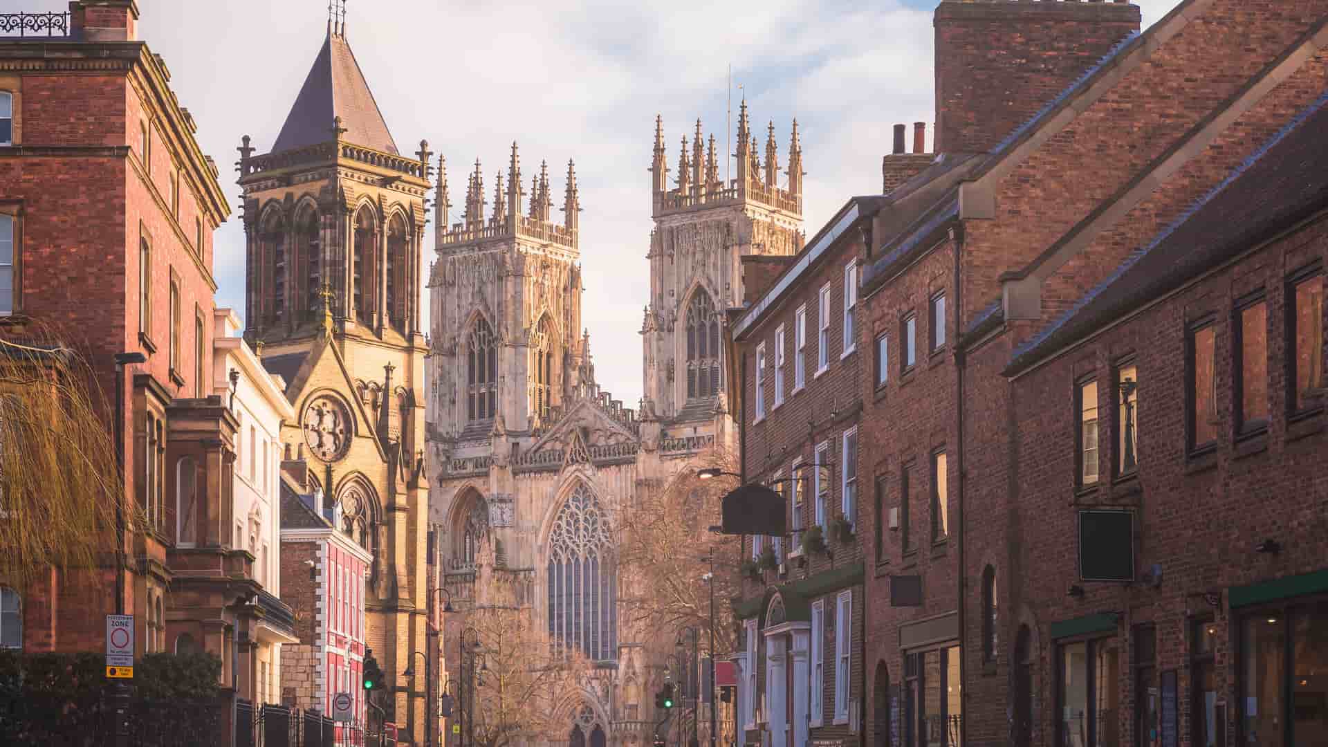 A historic street in York, North Yorkshire, England, with a prominent view of the towering spires of York Minster, the large gothic cathedral, flanked by traditional brick and stone buildings.