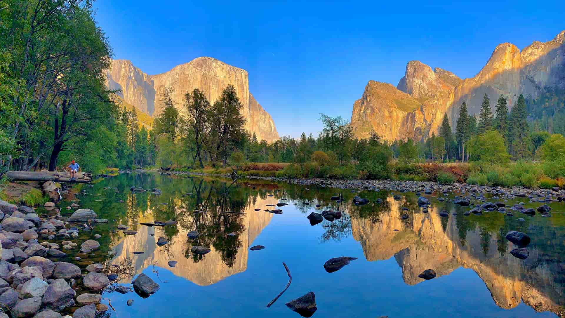 "A scenic panoramic shot of Yosemite Valley in California, with the iconic granite cliffs of El Capitan and Half Dome reflecting perfectly on the Merced River at sunset."
