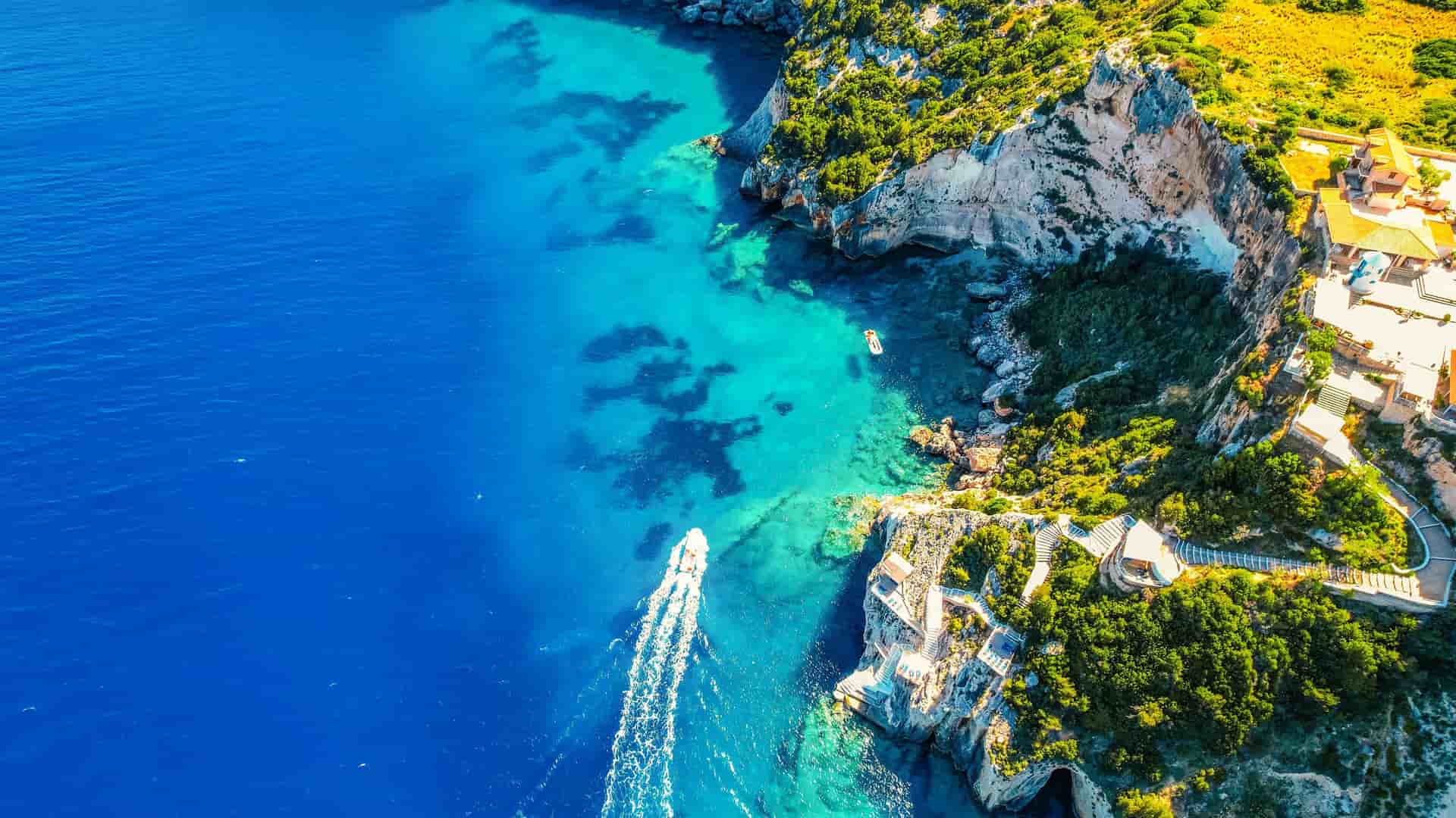 An aerial view of the vibrant blue waters and lush green cliffs of the Ionian Sea coastline in Zakynthos, Greece, with boats cruising offshore.