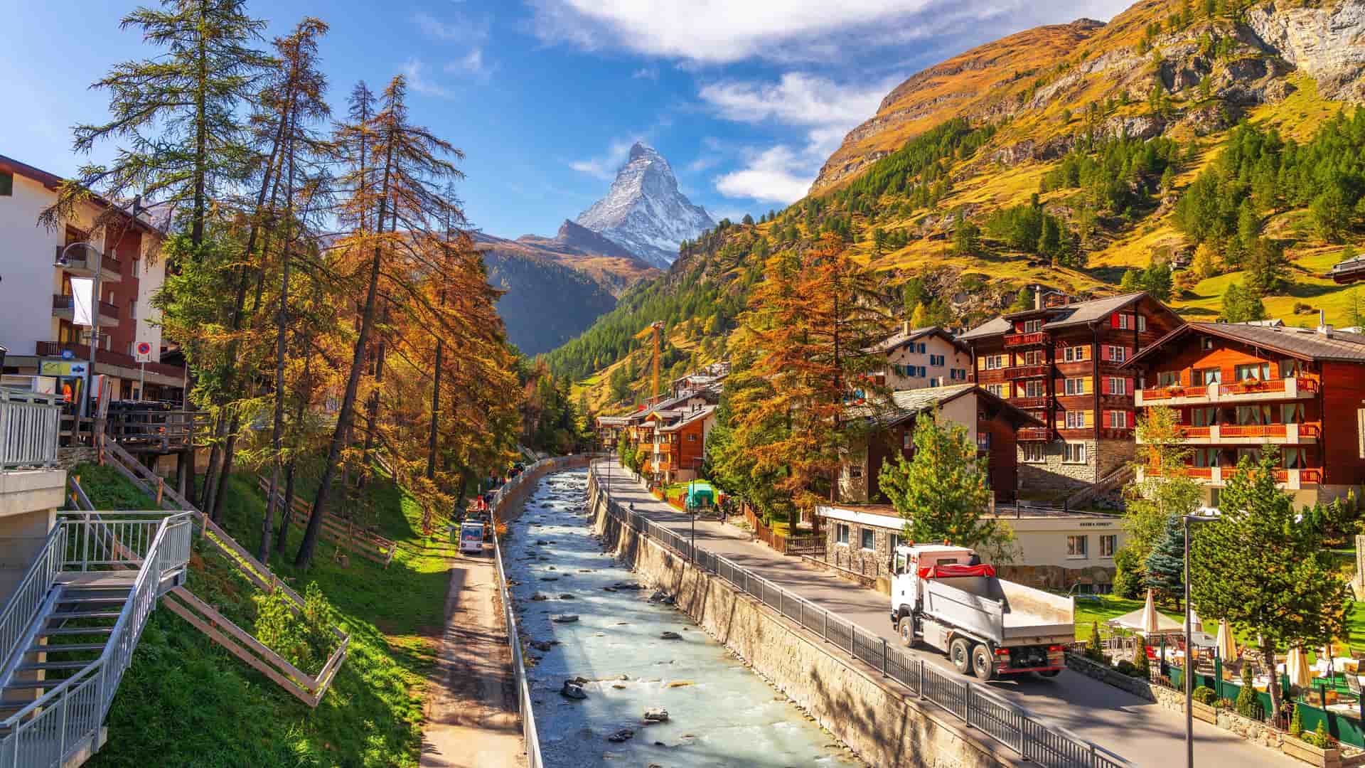 A vibrant autumn view of the Zermatt village in Switzerland, with the iconic Matterhorn mountain in the background, surrounded by colorful trees and a flowing river.
