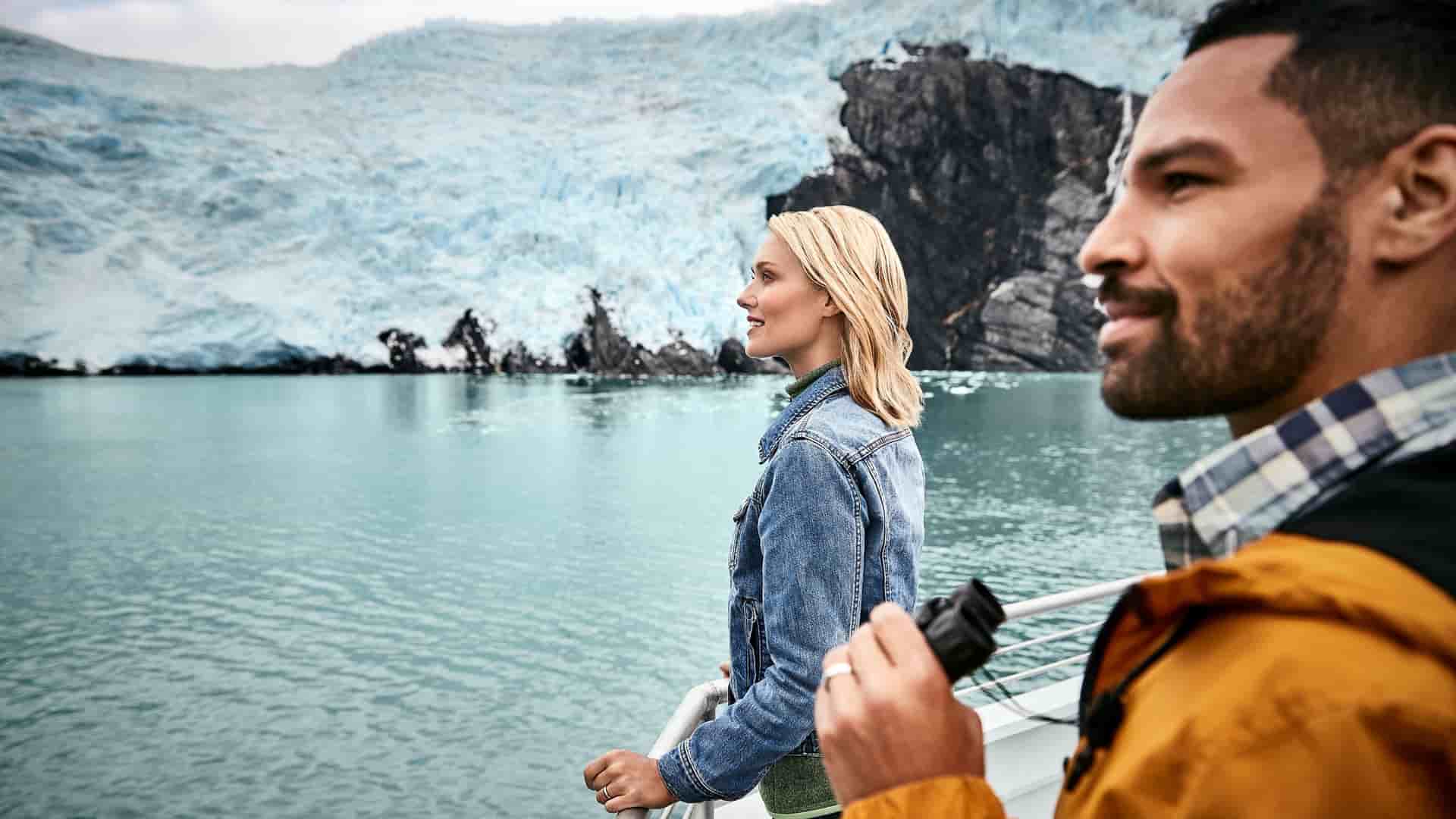 Two travelers stand on a boat deck looking at a massive blue glacier across calm water in Alaska.