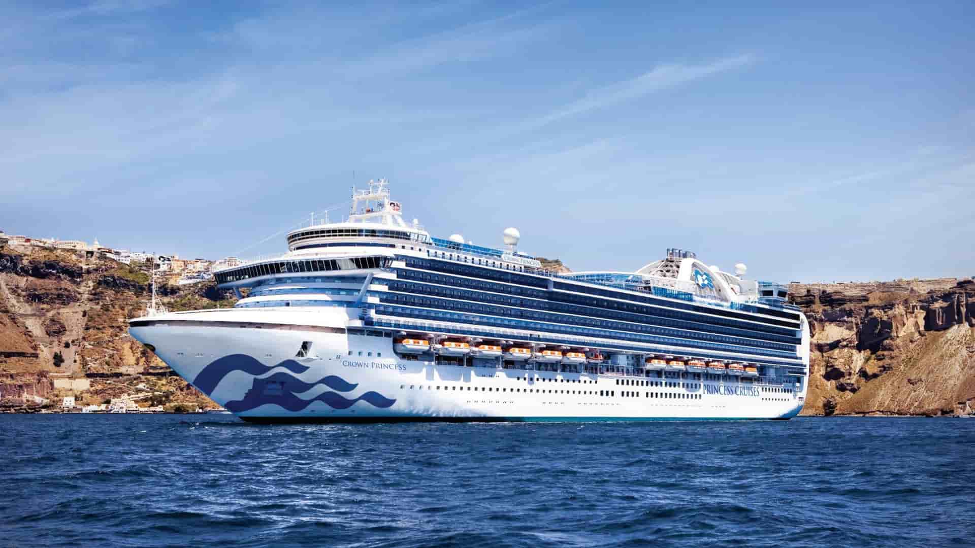 The Crown Princess cruise ship at sea, with the iconic white and blue buildings of Santorini, Greece visible on the cliffs in the background.