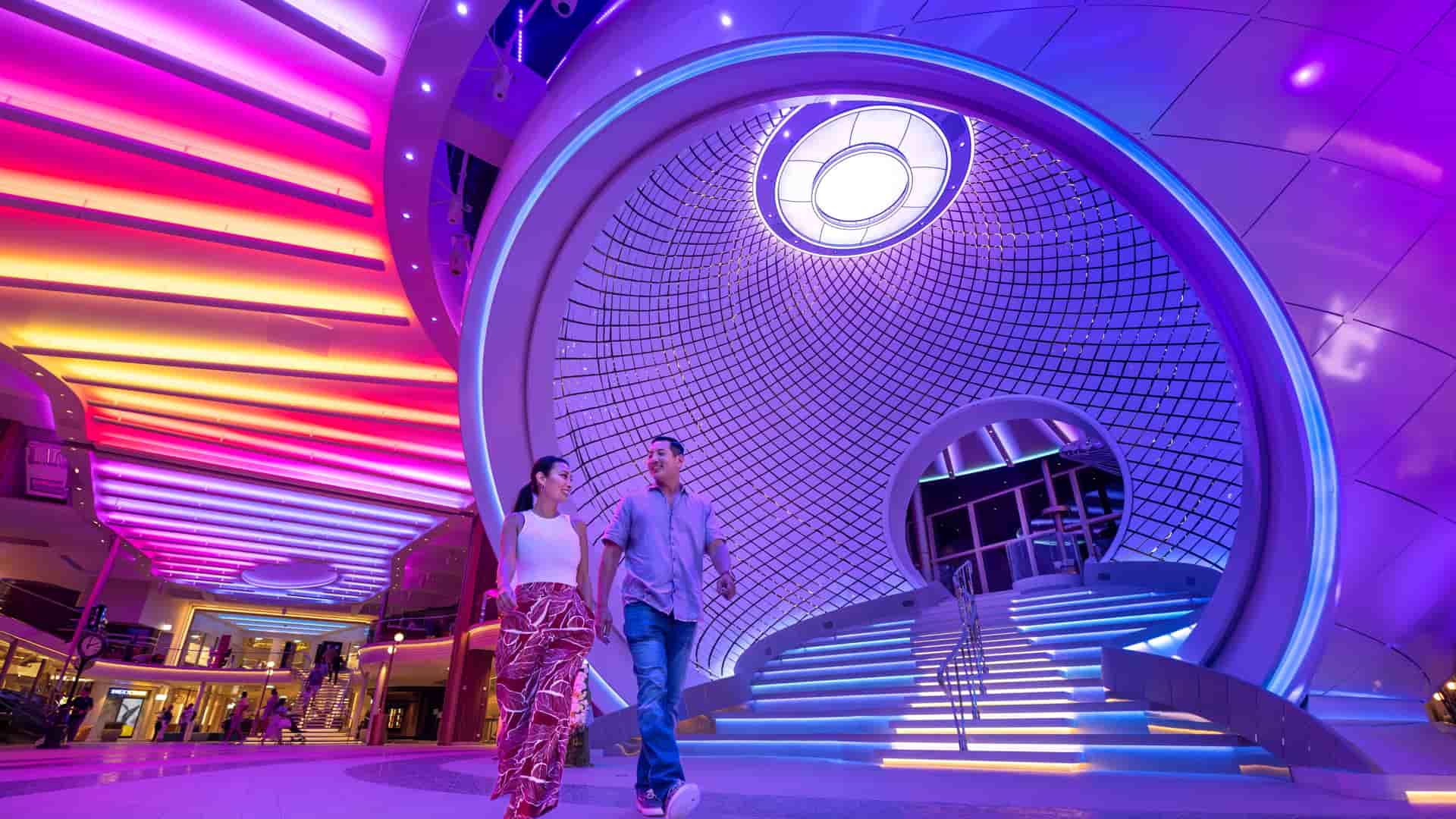 A straight couple walking in front of The Pearl, a three-deck-tall kinetic art installation that syncs to music, aboard Icon of the Seas.. 