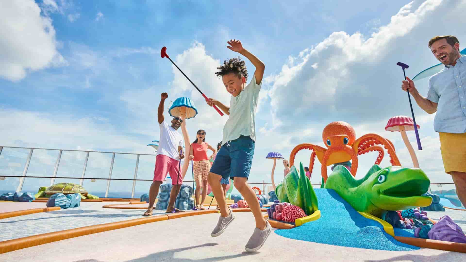 A young boy jumps for joy after sinking a mini-golf ball on the Royal Caribbean International Wonder of the Seas ship, with other people playing in the background.