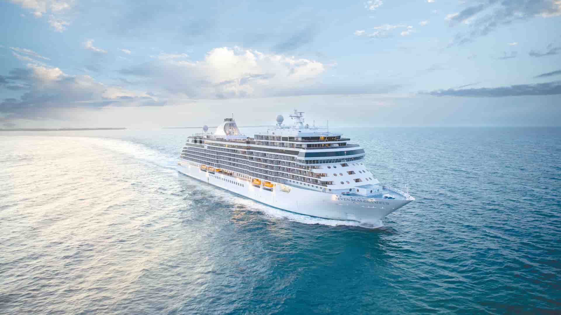 The luxury cruise ship Seven Seas Grandeur sails through the open blue ocean, leaving a white wake beneath a soft, cloudy sky.