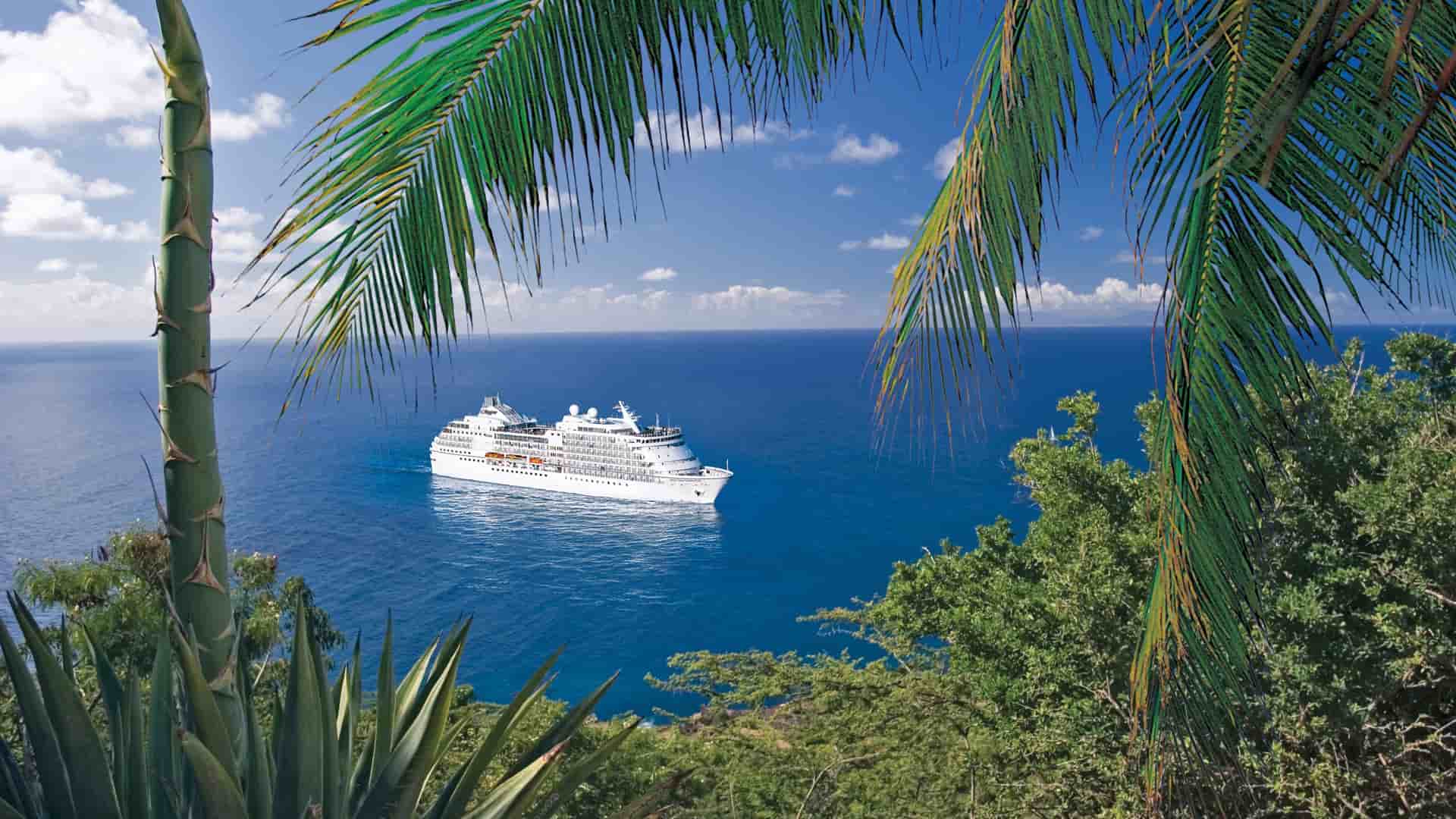 A luxury Regent Seven Seas cruise ship sailing across a deep blue ocean, framed by tropical palm trees and lush green foliage under a sunny sky.