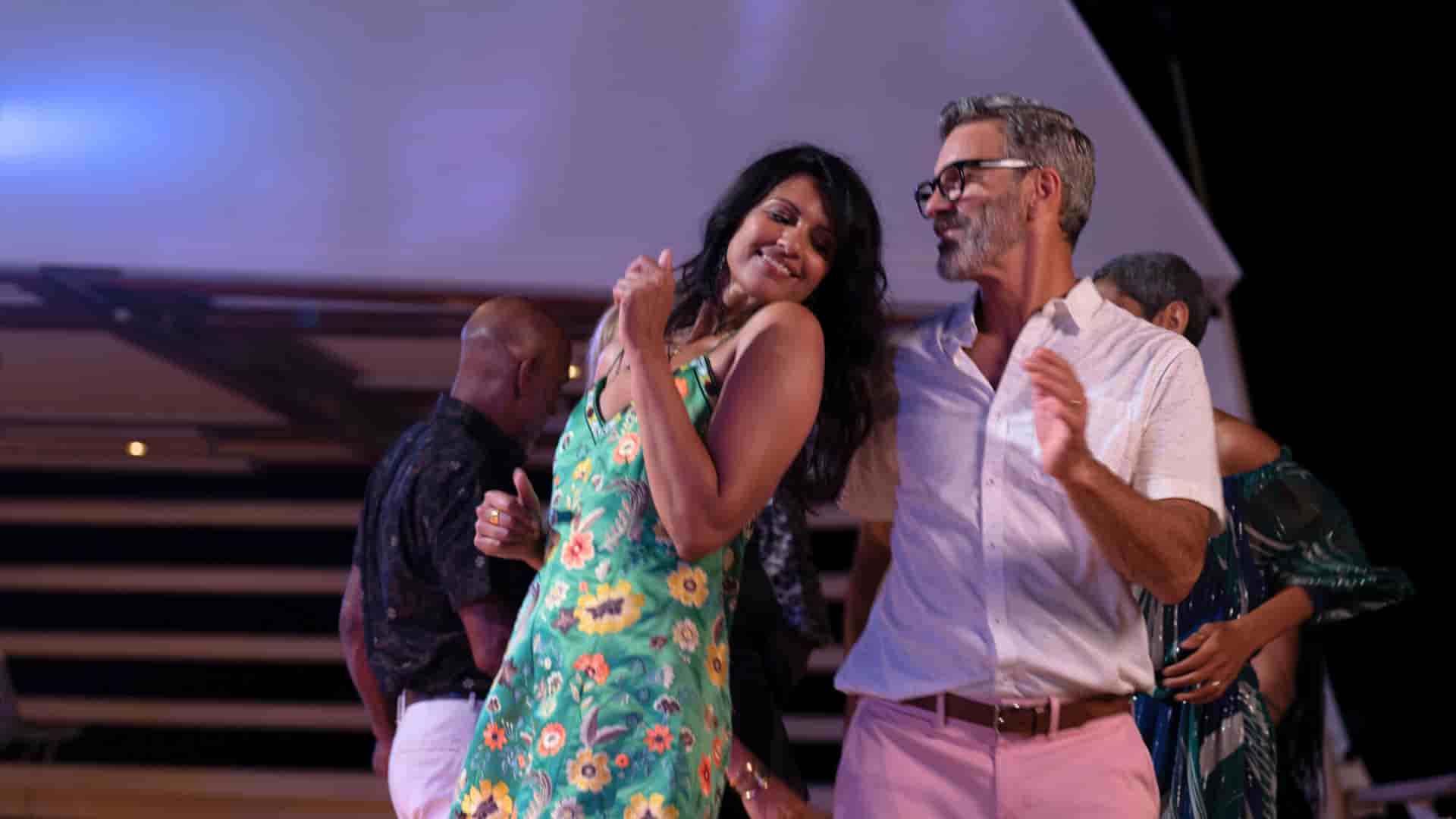 A man and woman are dancing together at night on the Seabourn Cruise Line's Seabourn Ovation ship, with the woman wearing a green floral dress and the man wearing a white shirt and pink pants.