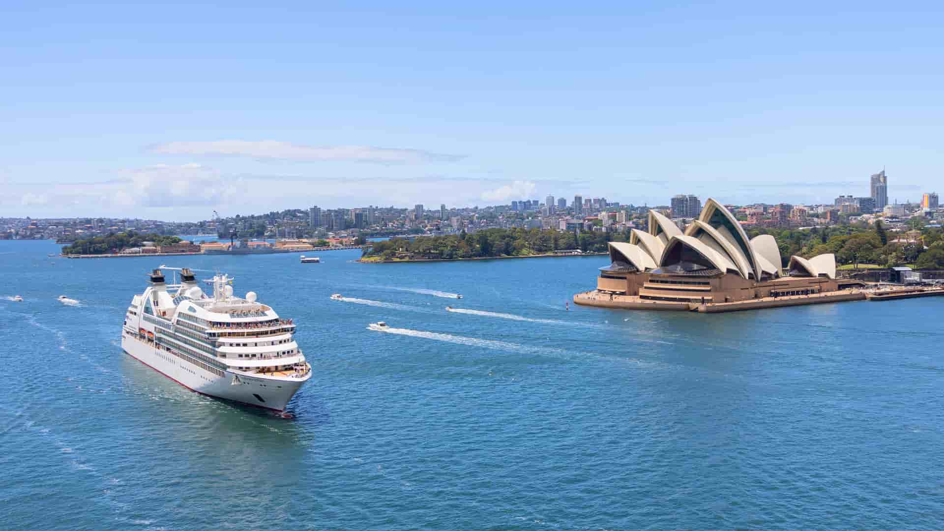 A Seabourn cruise ship sails past the iconic Sydney Opera House and city skyline in Australia, under a beautiful sky.