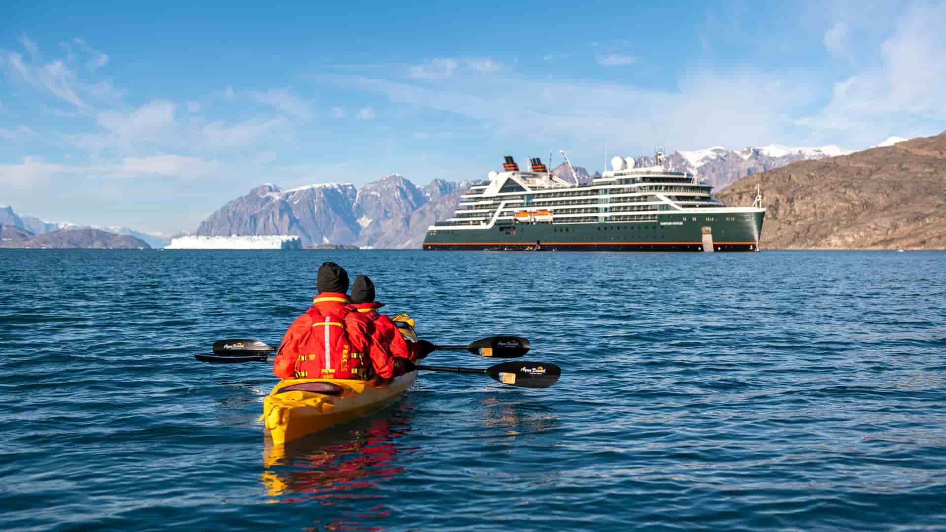 Two people kayak in the waters of Greenland with a Seabourn Venture cruise ship and snow-capped mountains in the background.
