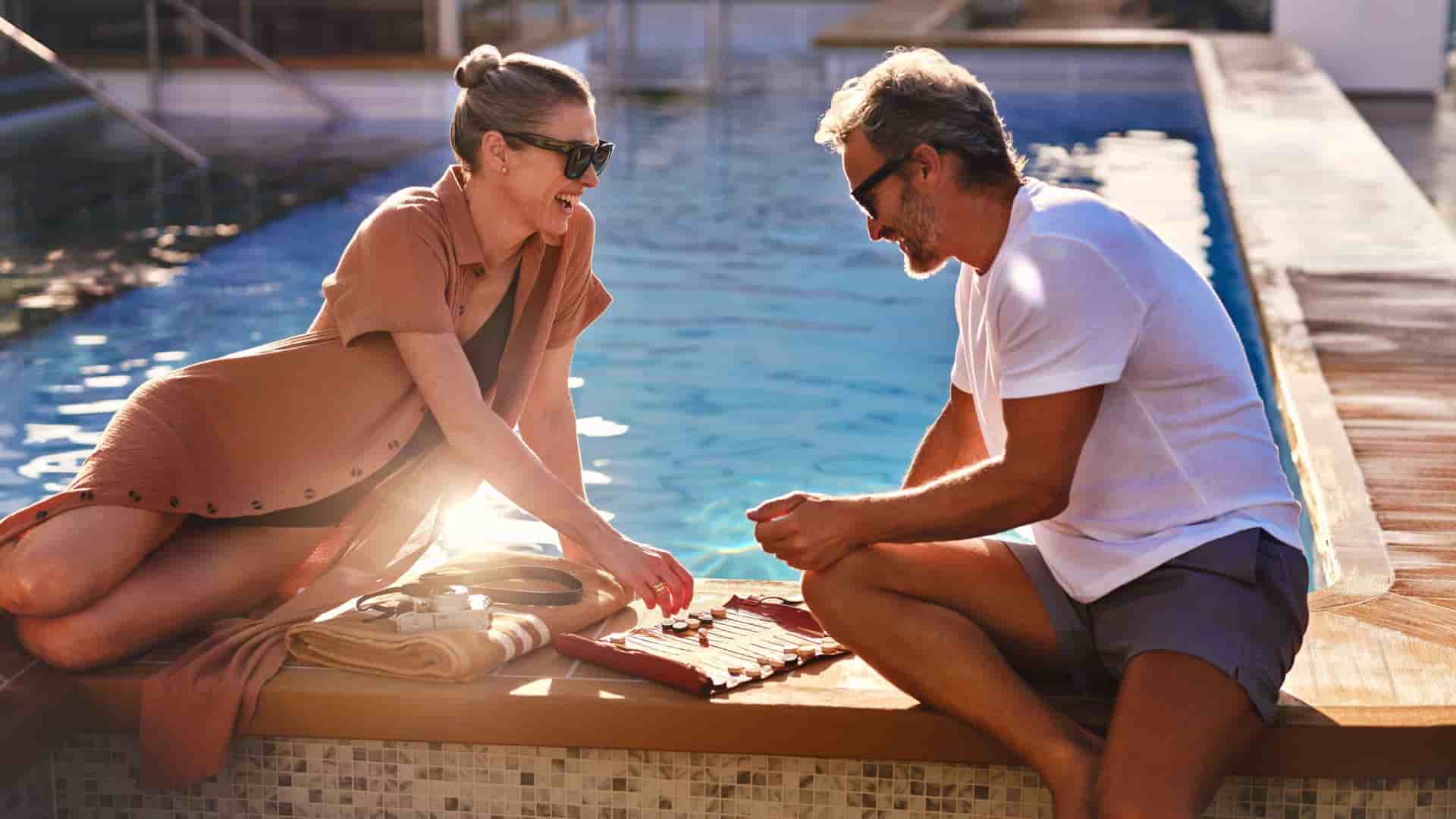 A happy couple on a Silversea cruise relaxes poolside, playing a game together on a sunny day.