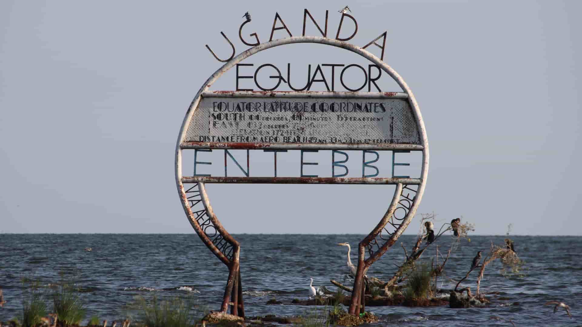 The Entebbe Equator marker standing in Lake Victoria, with rusted metal rings, perched birds, and calm water surrounding the landmark.