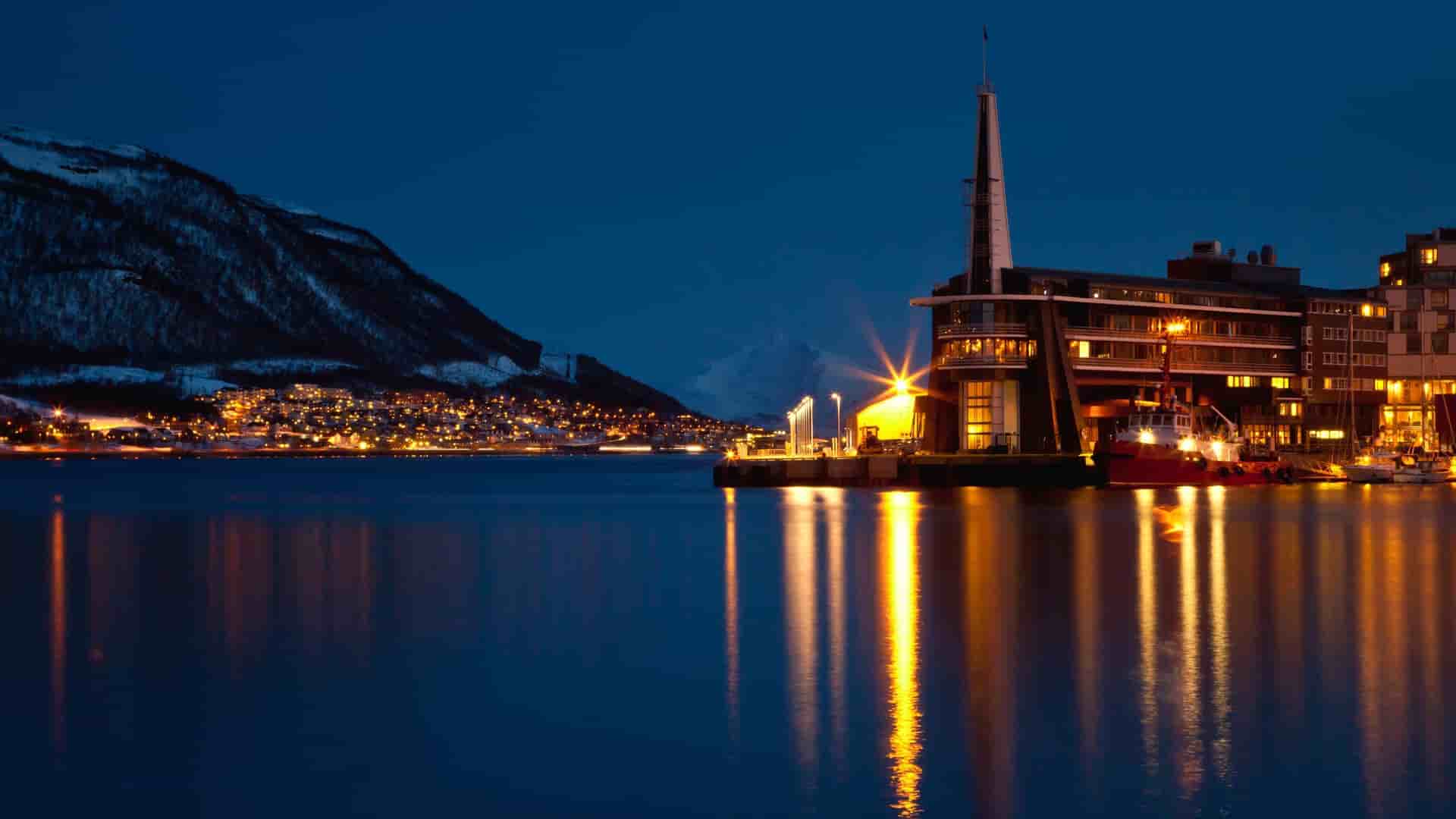 Tromsø harbor at night with illuminated buildings and snowy mountains.