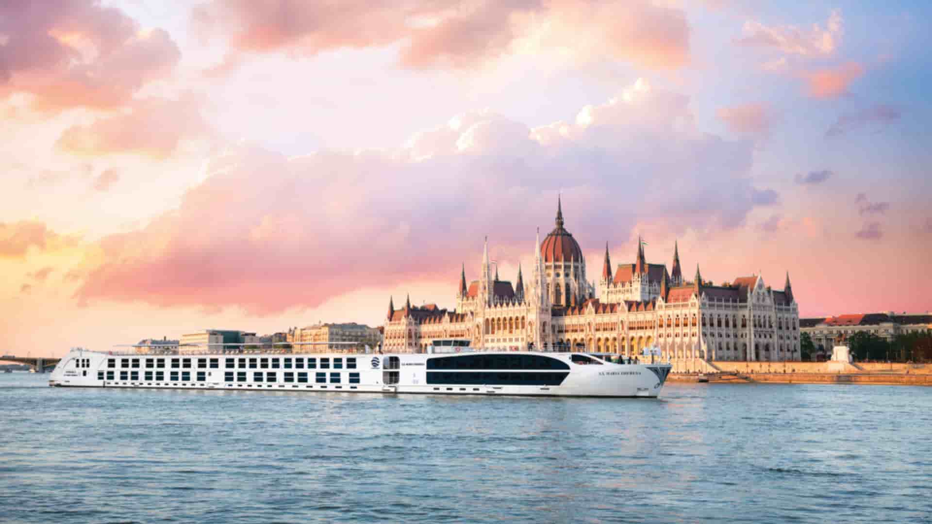 Uniworld's S.S. Maria Theresa river cruise ship sails on the Danube River with the Hungarian Parliament Building in Budapest, Hungary, under a dramatic pink and purple sky.