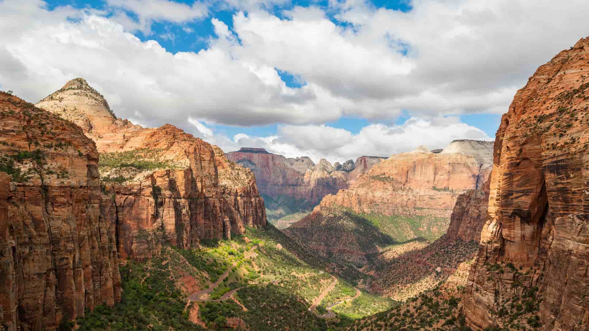 A sweeping aerial view of Zion National Park’s red rock canyons under a bright blue sky with fluffy white clouds. A winding road snakes through the lush green valley floor below, surrounded by massive sandstone cliffs and jagged mountain peaks.