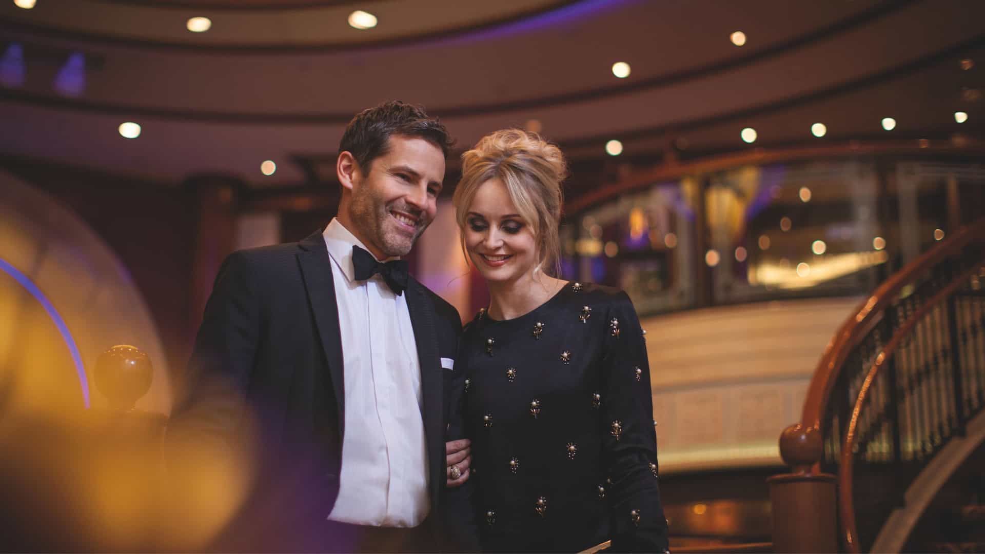 Couple smiling in formal attire as they walk through the Grand Lobby on a Cunard ship.