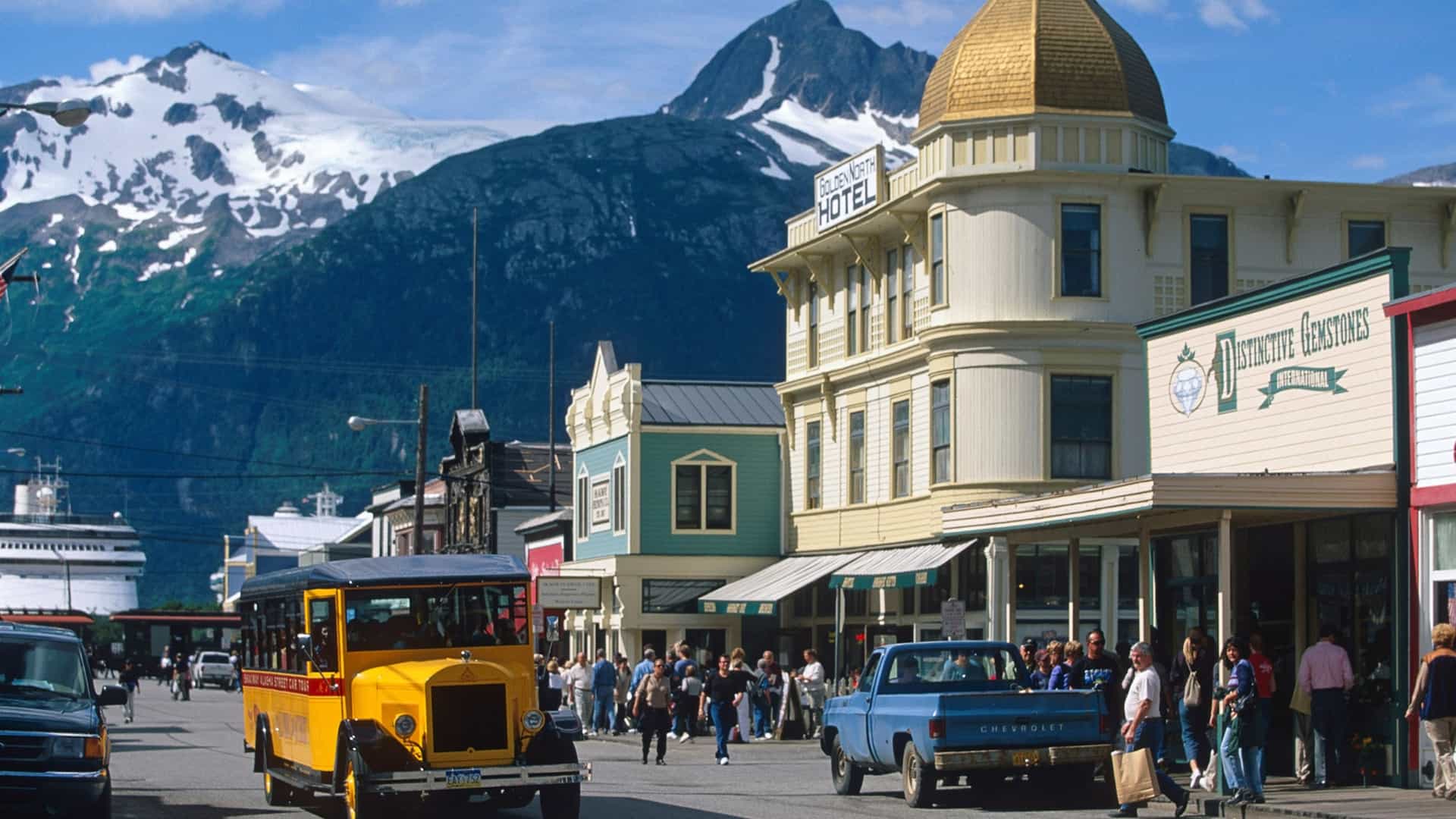 A vintage yellow tour bus and pedestrians travel down a historic street in Skagway, Alaska, lined with old-fashioned buildings against a backdrop of towering snow-capped mountains.