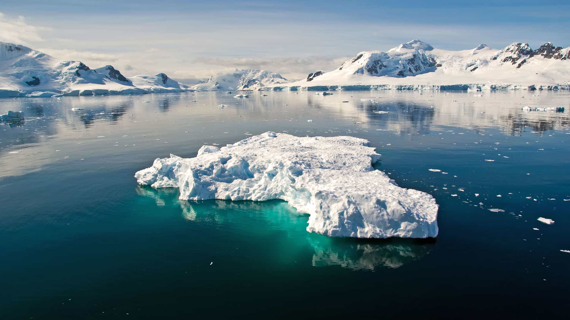 A large, flat iceberg with a bright turquoise base floats in the calm, blue waters of Antarctica. The iceberg's reflection is clearly visible on the glassy surface. In the background, majestic snow-covered mountains rise under a bright, partly cloudy sky. The scene is a striking depiction of the pristine, frozen landscape and vastness of the Antarctic region.