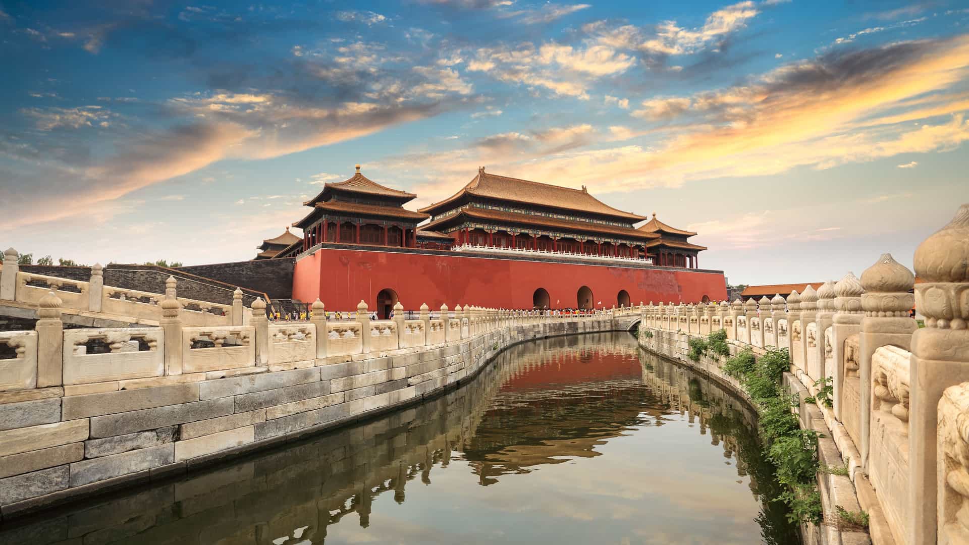 The iconic red walls and golden roofs of the Forbidden City in Beijing, China, are beautifully reflected in the calm waters of the surrounding moat. A traditional stone bridge with ornate carvings crosses the water, leading toward the grand palace buildings. The sky above is a vibrant mix of blue, orange, and white clouds during sunset. The image captures the historical grandeur and imperial architecture of this significant landmark in the Asian region.