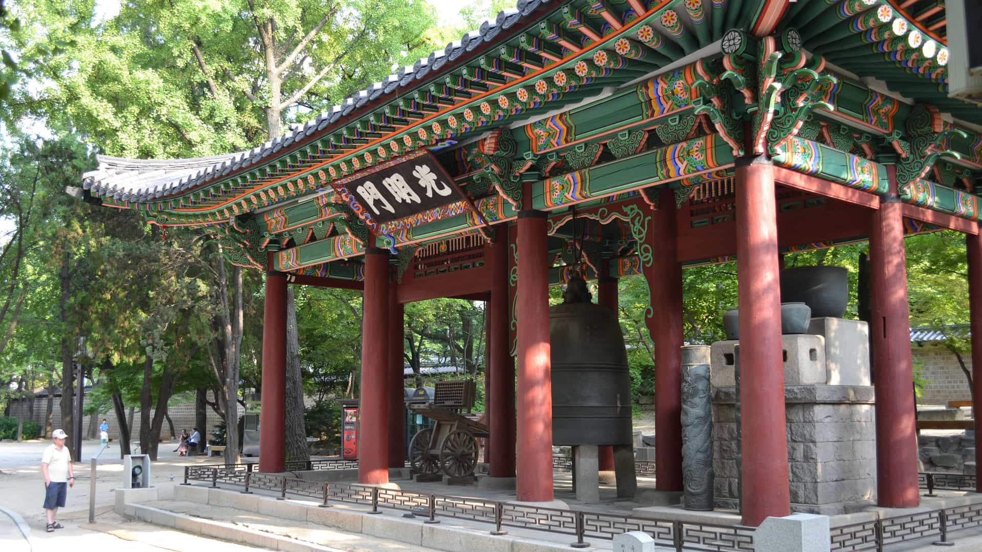 A traditional Korean bell pavilion within a temple in Seoul, Korea. The pavilion has a classic sloping roof with intricate, colorful patterns in green, red, and orange, supported by thick red wooden pillars. A large bronze bell hangs inside the structure, next to a stone pedestal. The setting is surrounded by lush green trees and a well-maintained stone courtyard. The image captures the unique architecture and peaceful atmosphere of a cultural landmark in the Asian region.