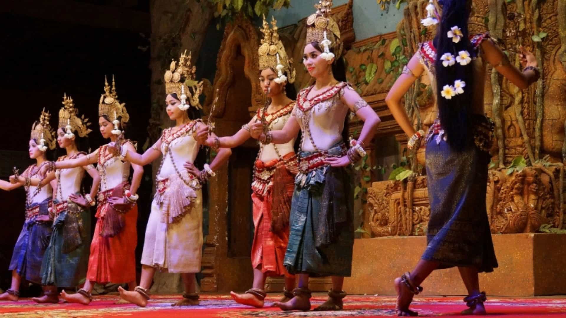 A group of women in vibrant traditional costumes perform an Apsara dance on a stage in Cambodia. The dancers wear elaborate golden headdresses, silk skirts in various colors, and ornate jewelry. Their hands are gracefully posed in traditional mudras, and they are adorned with fresh flowers in their hair. The performance space has intricately carved wooden backdrops, showcasing a cultural and artistic tradition of the Southeast Asian river region.