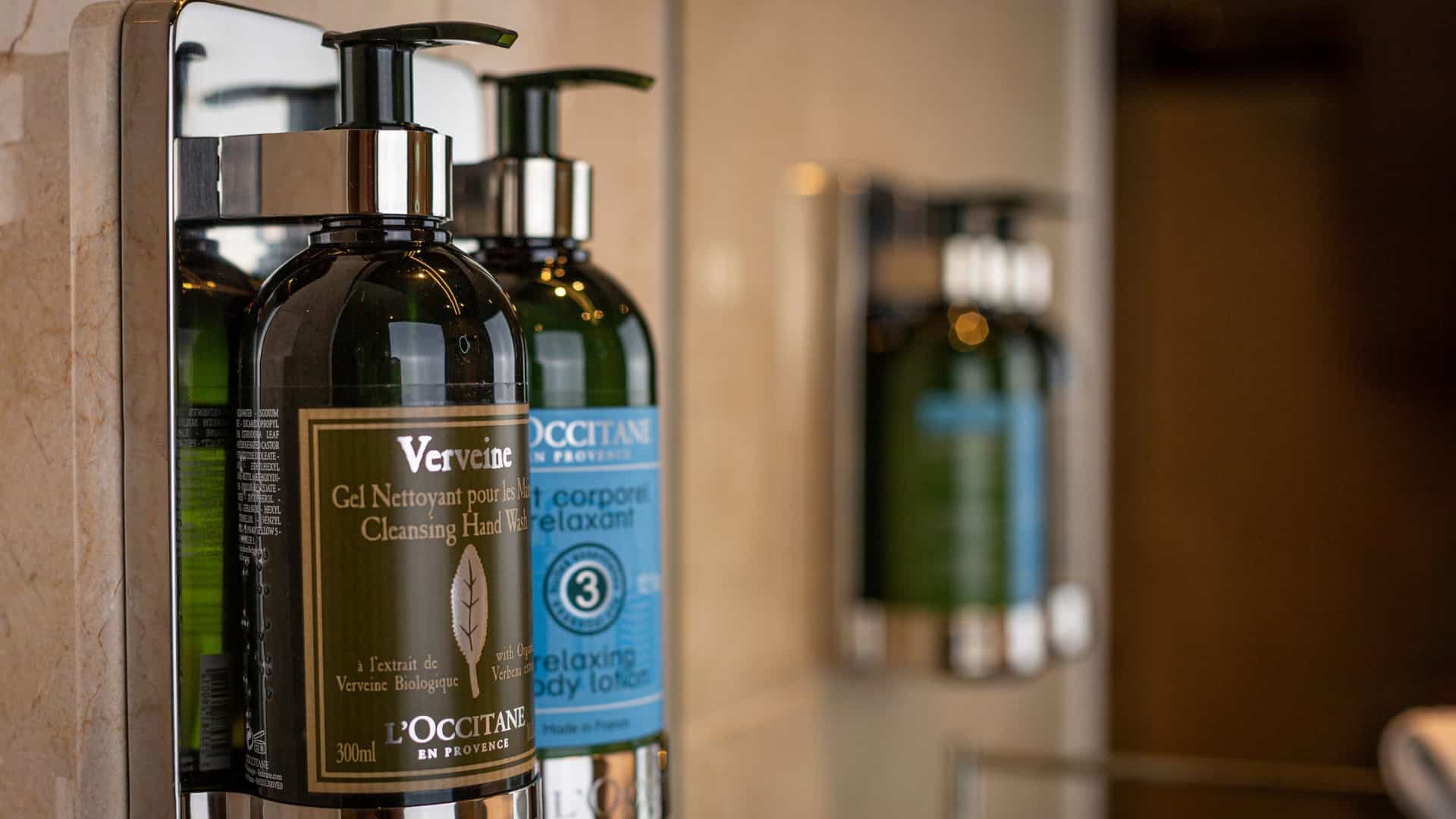 L'Occitane en Provence Verveine cleansing hand wash and body lotion pump dispensers mounted on a tiled bathroom wall.