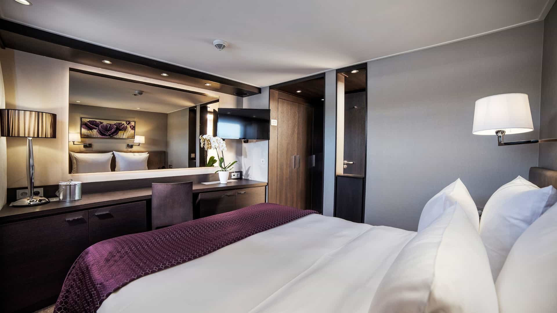 Deluxe stateroom aboard an Avalon Waterways river cruise ship featuring a bed with a deep purple runner, a large illuminated vanity mirror, a wooden desk with an orchid, and modern cabinetry.