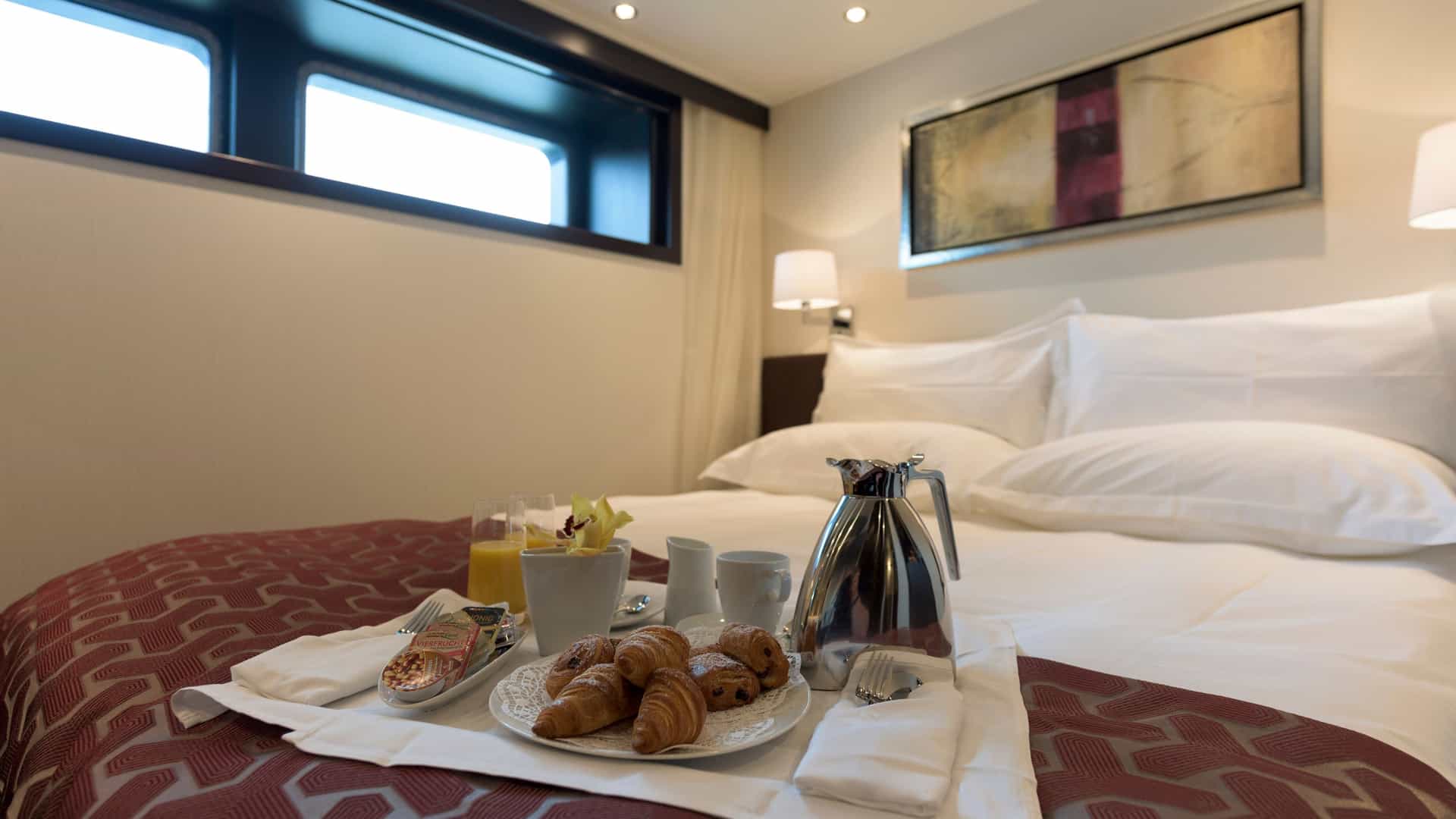 Continental breakfast tray with pastries, coffee, and orange juice resting on a bed with crisp white linens and a maroon patterned runner inside an Avalon Waterways river cruise deluxe stateroom.