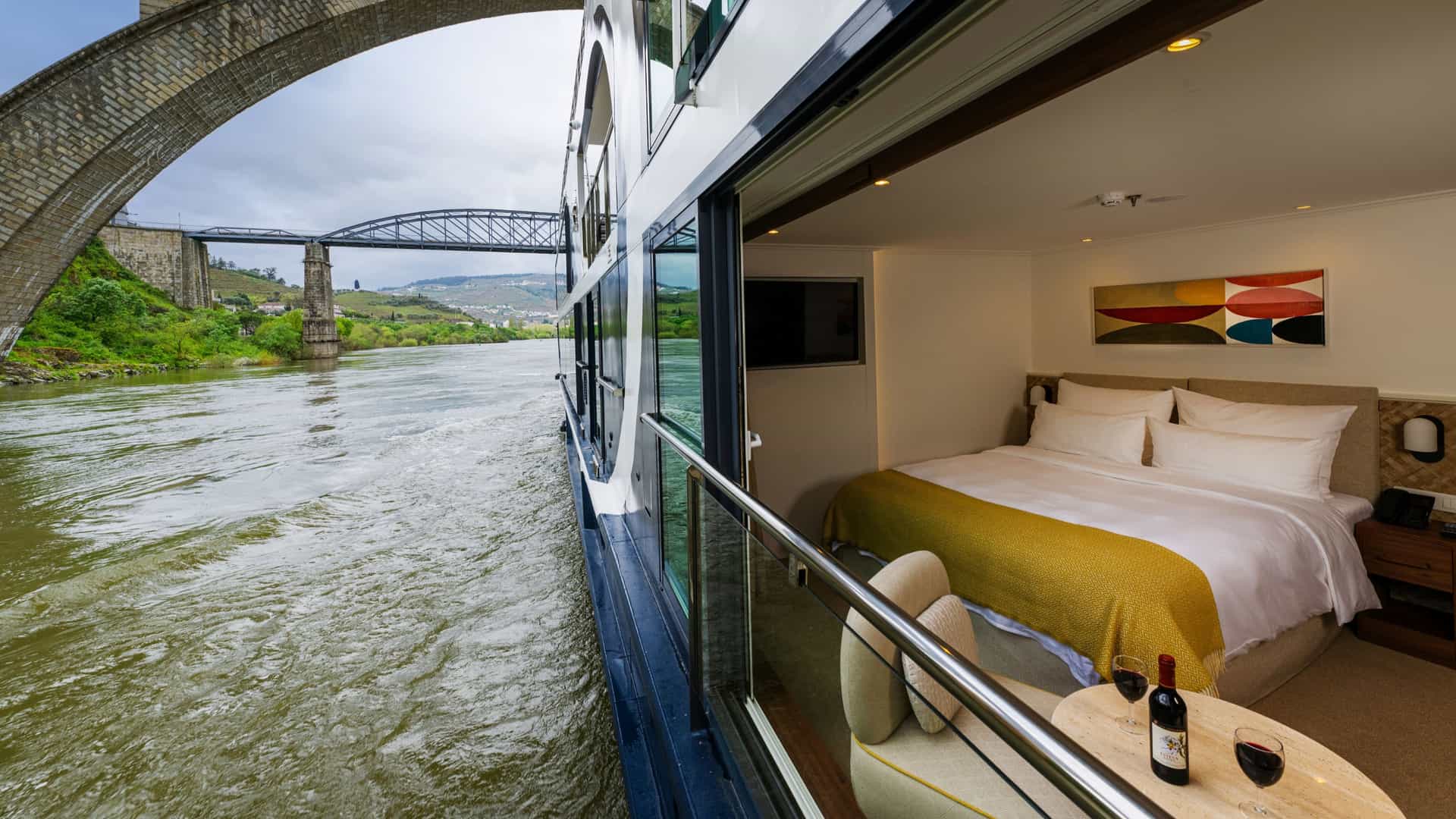 An open-air Panorama Suite aboard an Avalon Waterways ship featuring a bed facing a wide-open floor-to-ceiling glass wall, a small table with a wine bottle and two glasses, and a scenic view of a historic stone bridge and passing river.