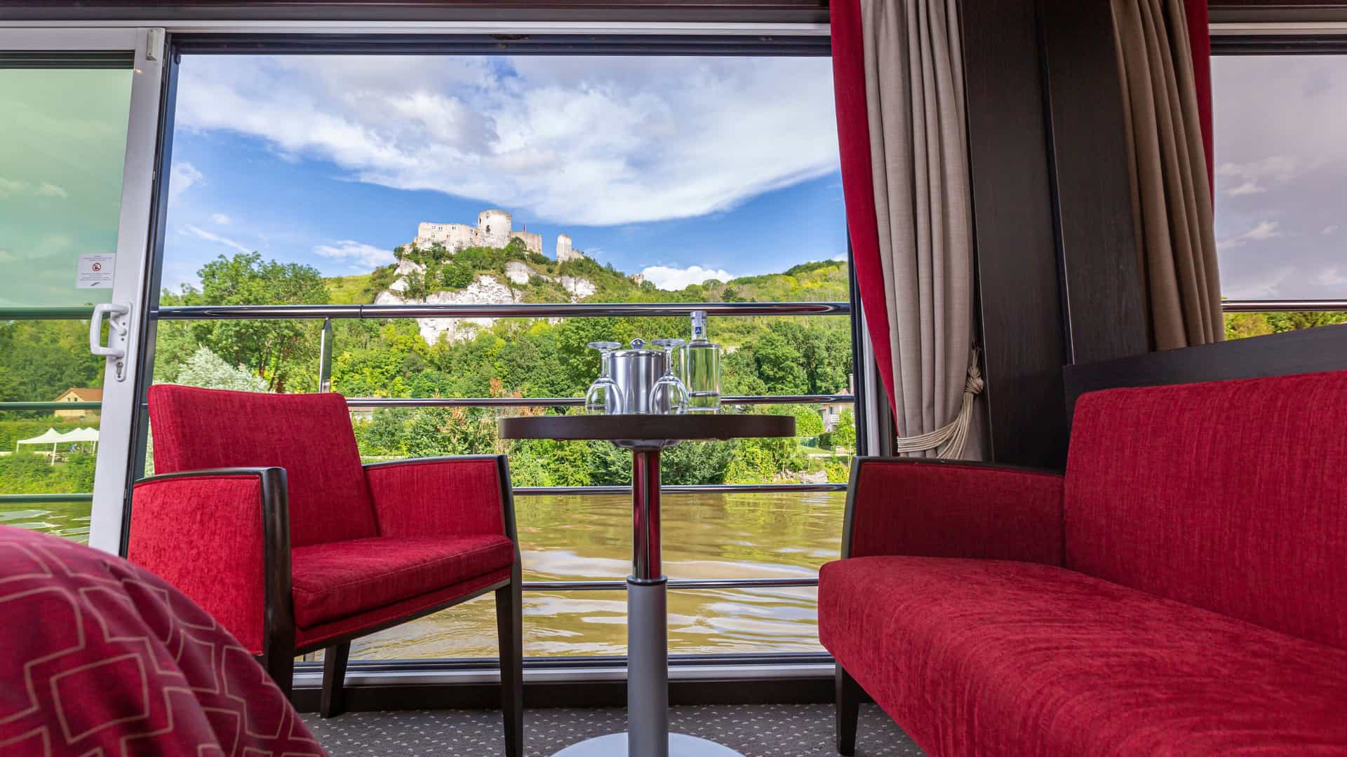Floor-to-ceiling panoramic sliding glass windows in an Avalon Waterways Panorama Suite, featuring red armchairs and a small table framing a scenic view of a river and a historic hilltop castle.