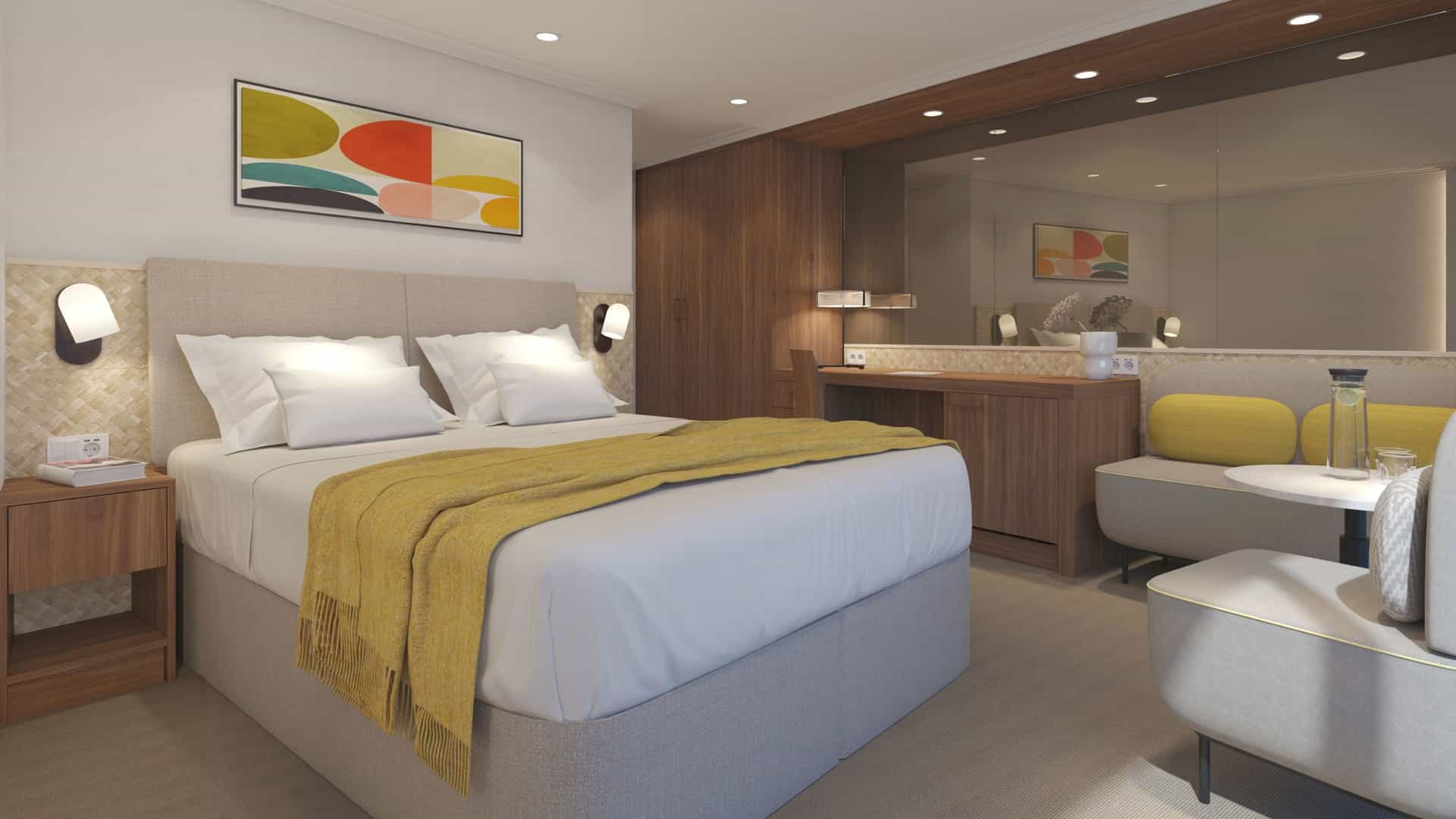 Modern suite interior on the Avalon Alegria featuring a plush bed with a mustard-yellow throw blanket, an abstract painting, a cozy seating area with yellow cushions, and a wooden vanity with a large mirror.