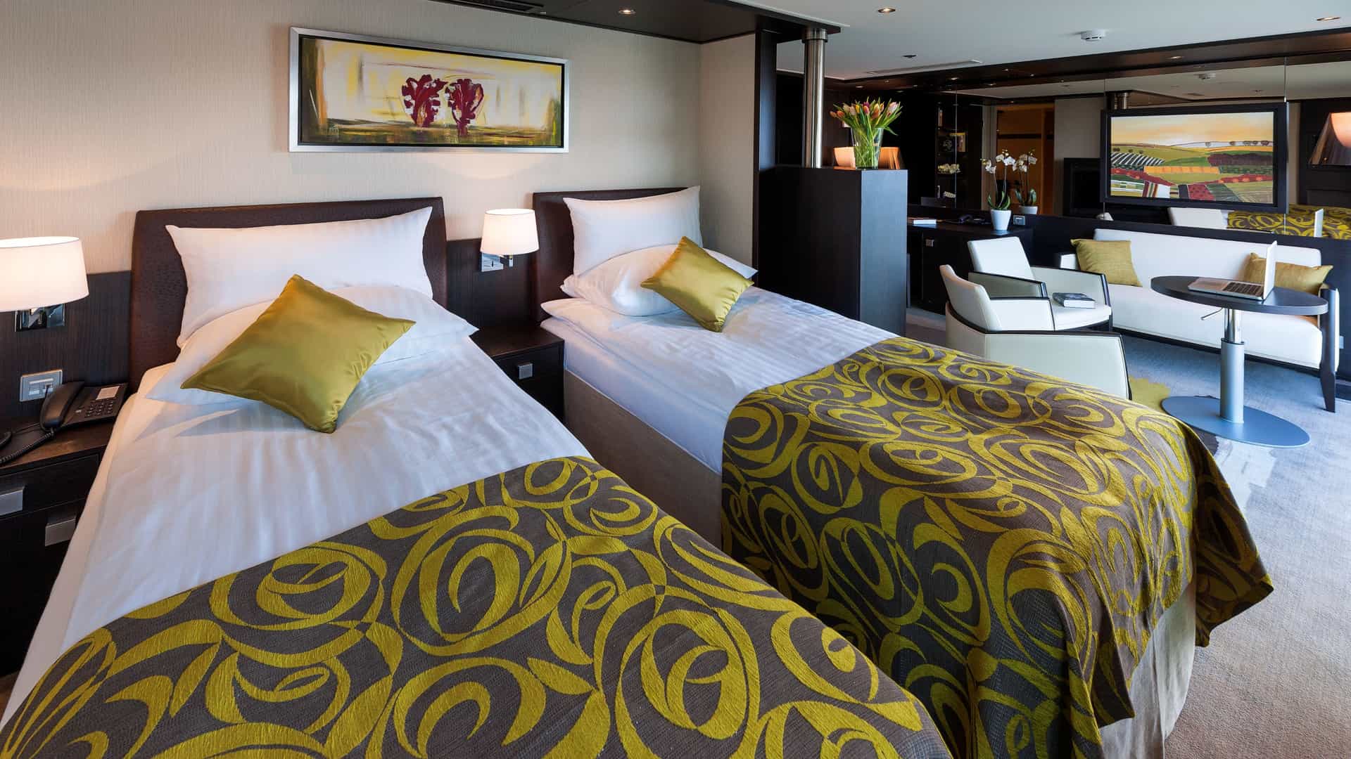 Spacious Royal Suite aboard an Avalon Waterways river cruise ship configured with two single beds featuring green and gold patterned runners and olive-green pillows, with an adjacent living area featuring a white sofa and armchairs.