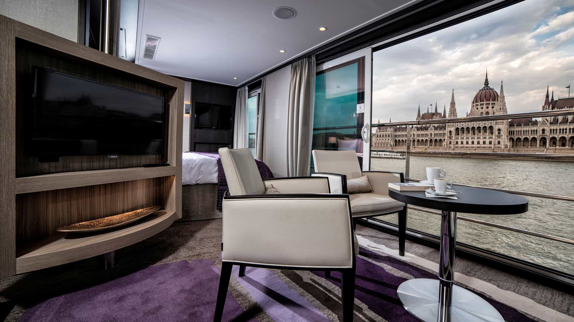 Floor-to-ceiling sliding glass windows in an Avalon Waterways Royal Suite, featuring cream armchairs and a small table framing a magnificent view of the Hungarian Parliament Building across the river.