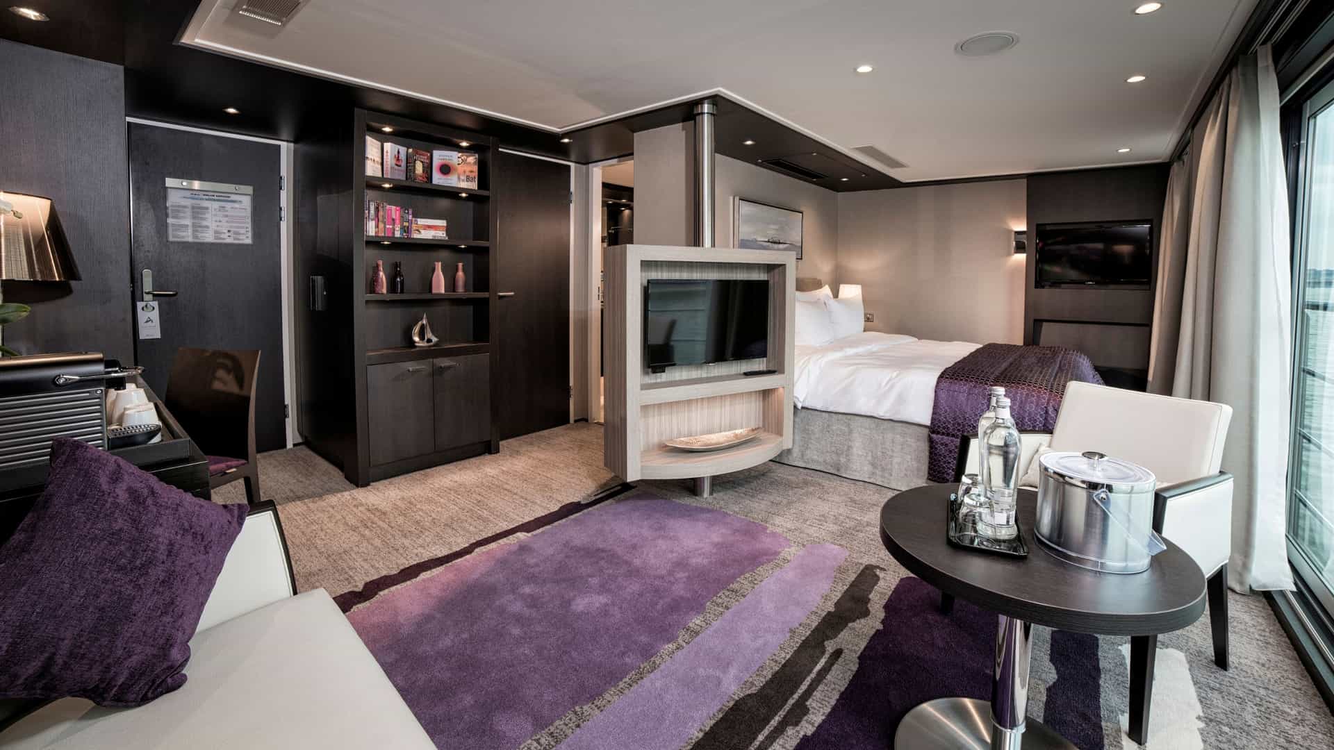 Spacious Royal Suite aboard an Avalon Waterways river cruise ship featuring a living area with a purple abstract rug, a plush bed with a deep purple runner, a central dividing wall with a mounted flat-screen TV, a built-in dark wood bookcase, and floor-to-ceiling sliding glass doors.