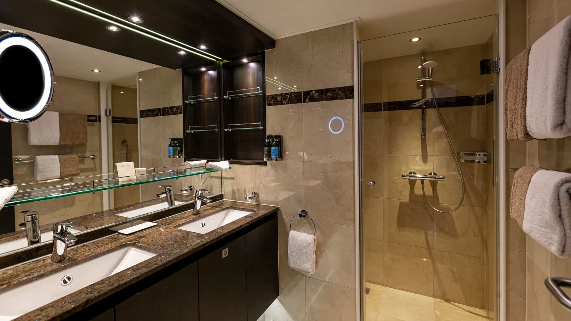 Modern bathroom in an Avalon Waterways Royal Suite featuring a double-sink vanity with dark cabinetry and a brown granite countertop, a large illuminated wall mirror, an adjustable makeup mirror, and a glass-enclosed walk-in shower with neutral tile work.